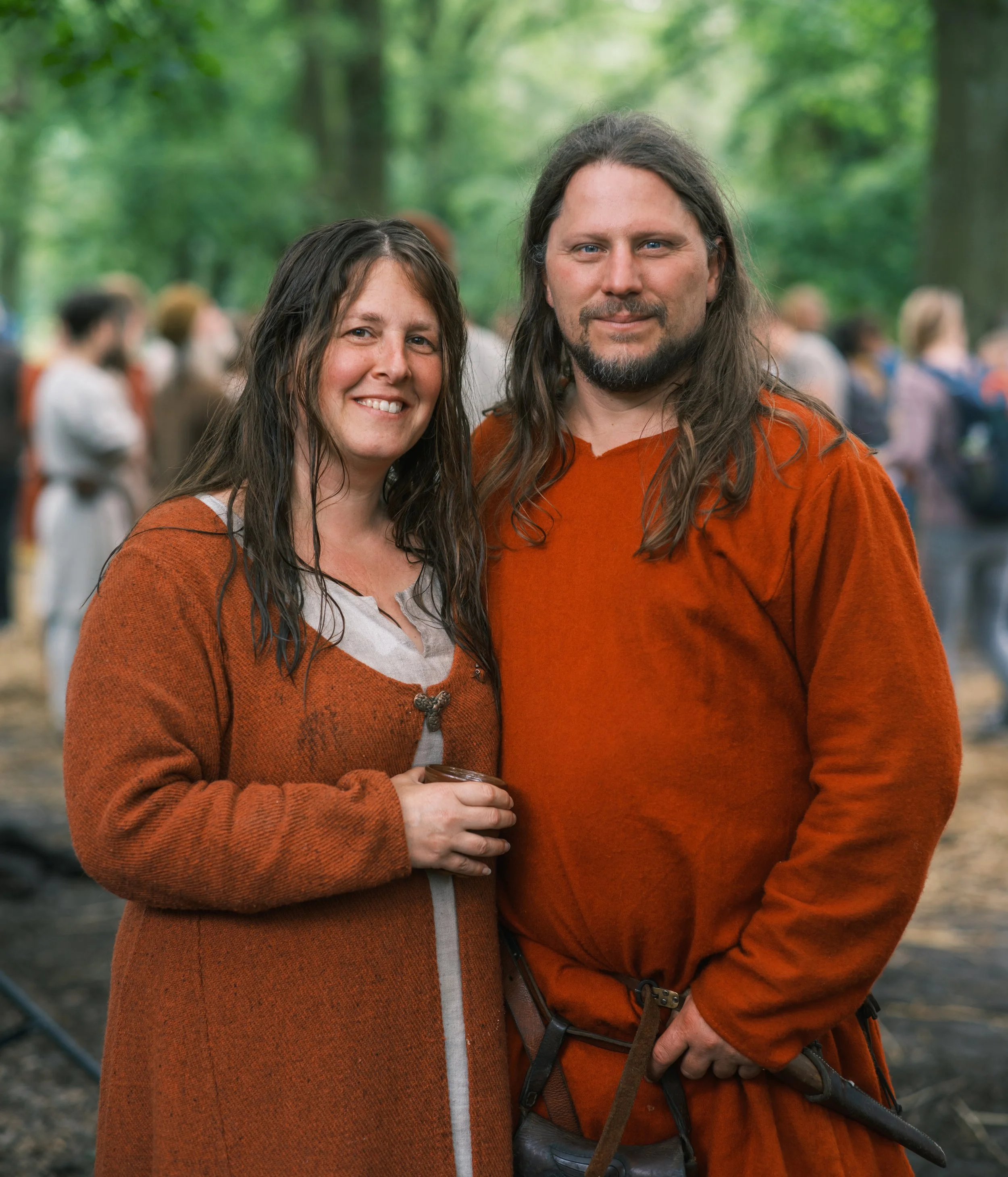 A woman and a man standing close together outdoors in a wooded area, smiling, with blurred people in the background.