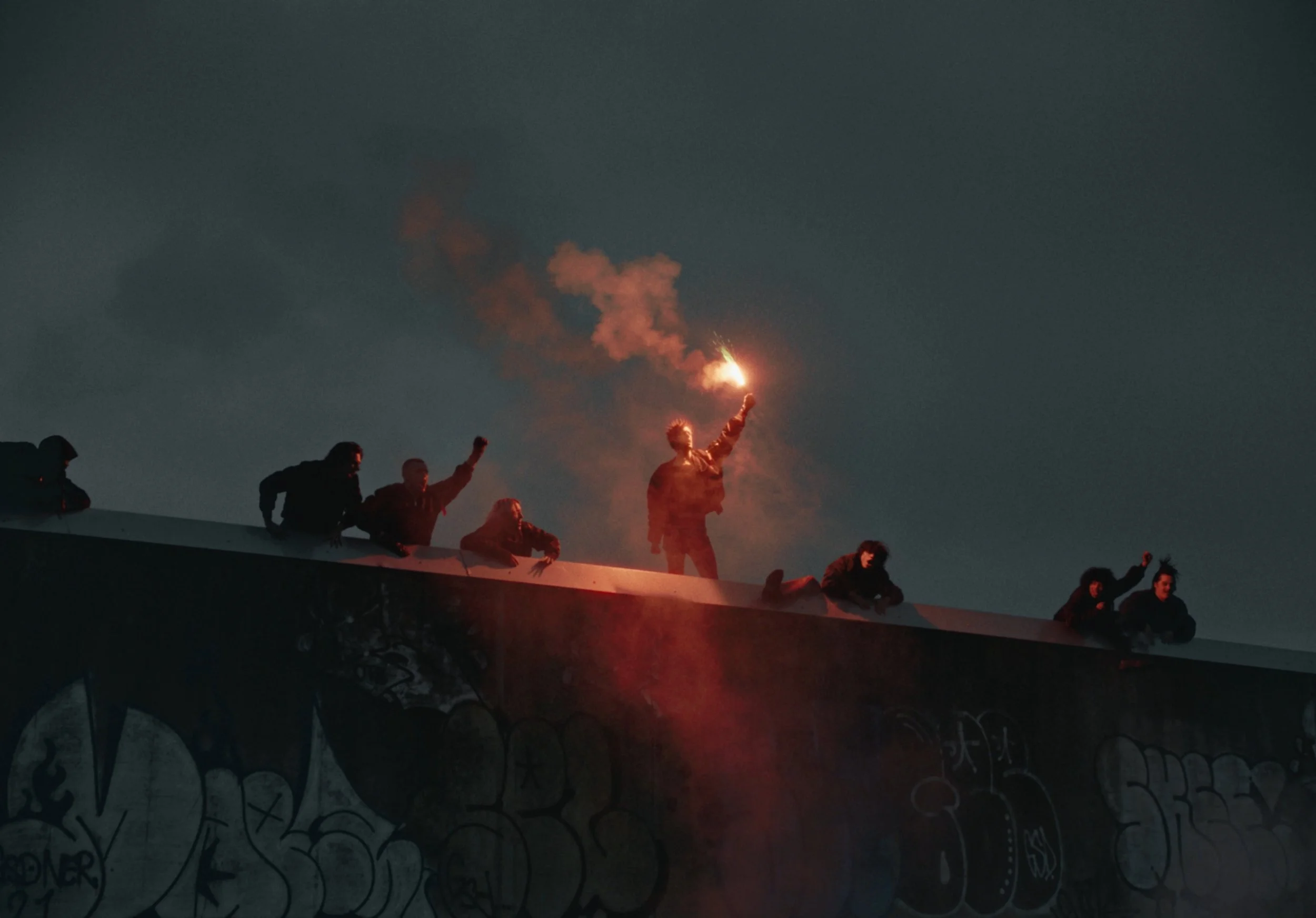 People standing on top of a structure at night, some holding a lit flare emitting orange smoke.