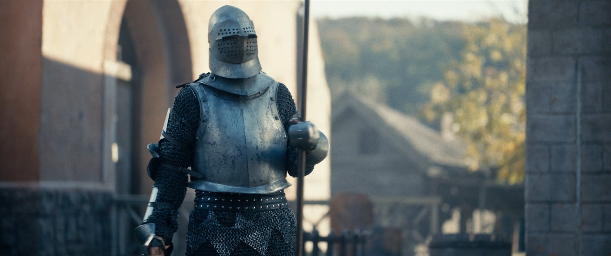 A person dressed as a medieval knight in full armor, holding a pole weapon, walking outdoors near stone walls and a bridge.