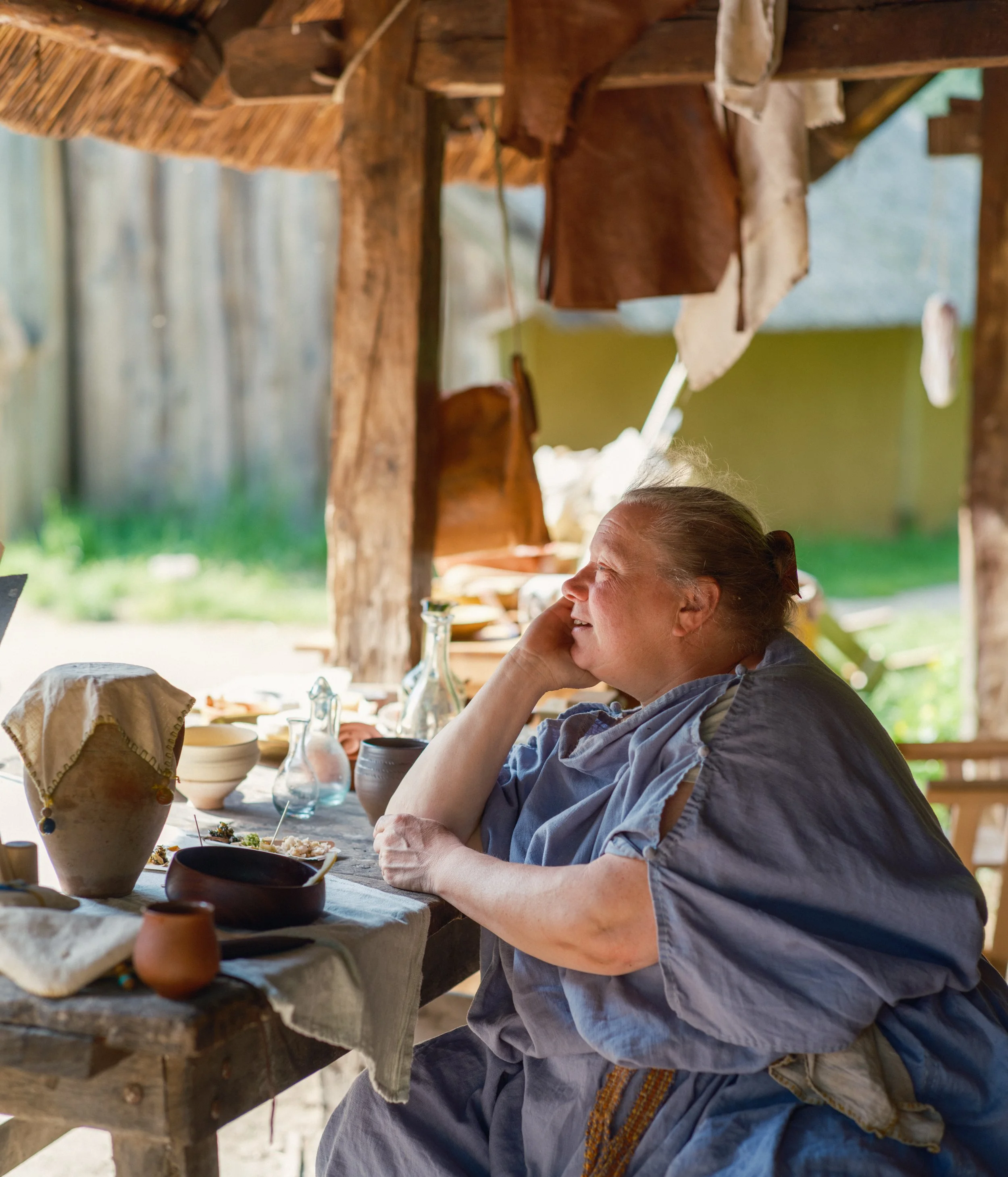 A woman with gray hair sitting at a rustic wooden table outdoors under a thatched roof. She is looking to her right, resting her head on her hand, and appears to be smiling. On the table, there are various ceramic dishes, a glass jar, and linen cloth