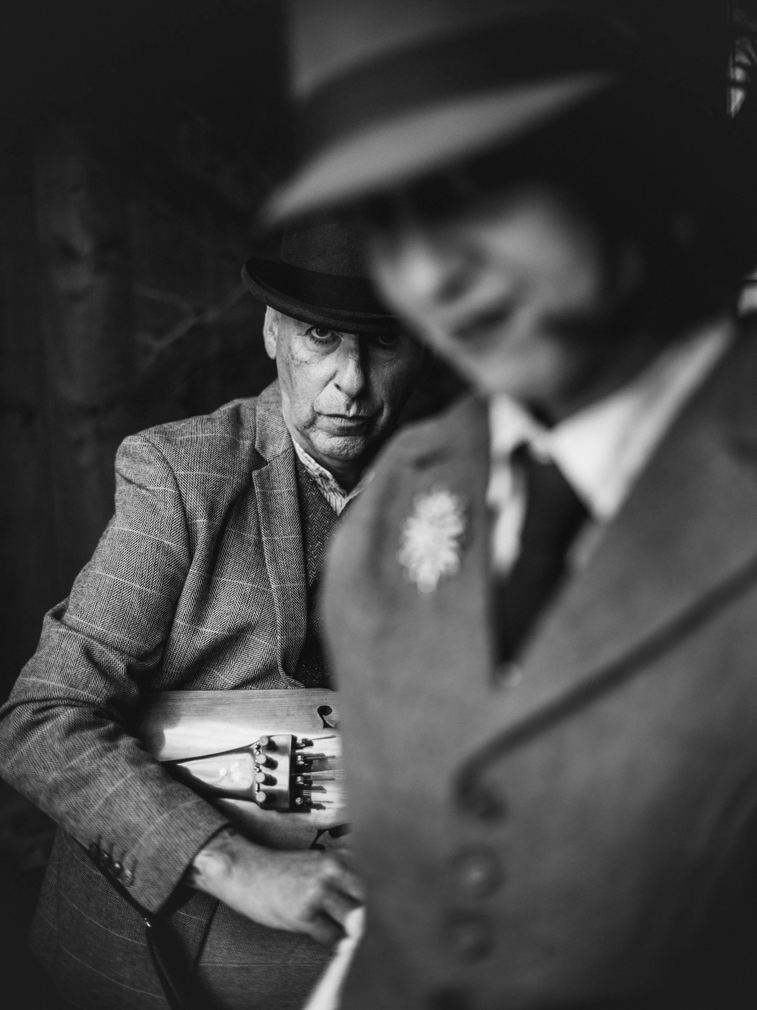 Black and white photo of a man wearing a hat and a suit, sitting at a table with a guitar, with another person in the foreground partially obscuring the view.