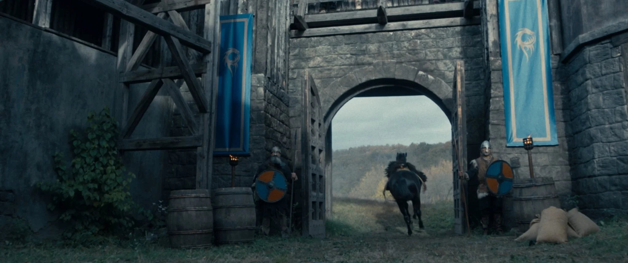 A medieval stone castle gate with two guards holding shields, a knight on horseback riding out, and blue banners with a dragon emblem hanging on both sides of the archway.