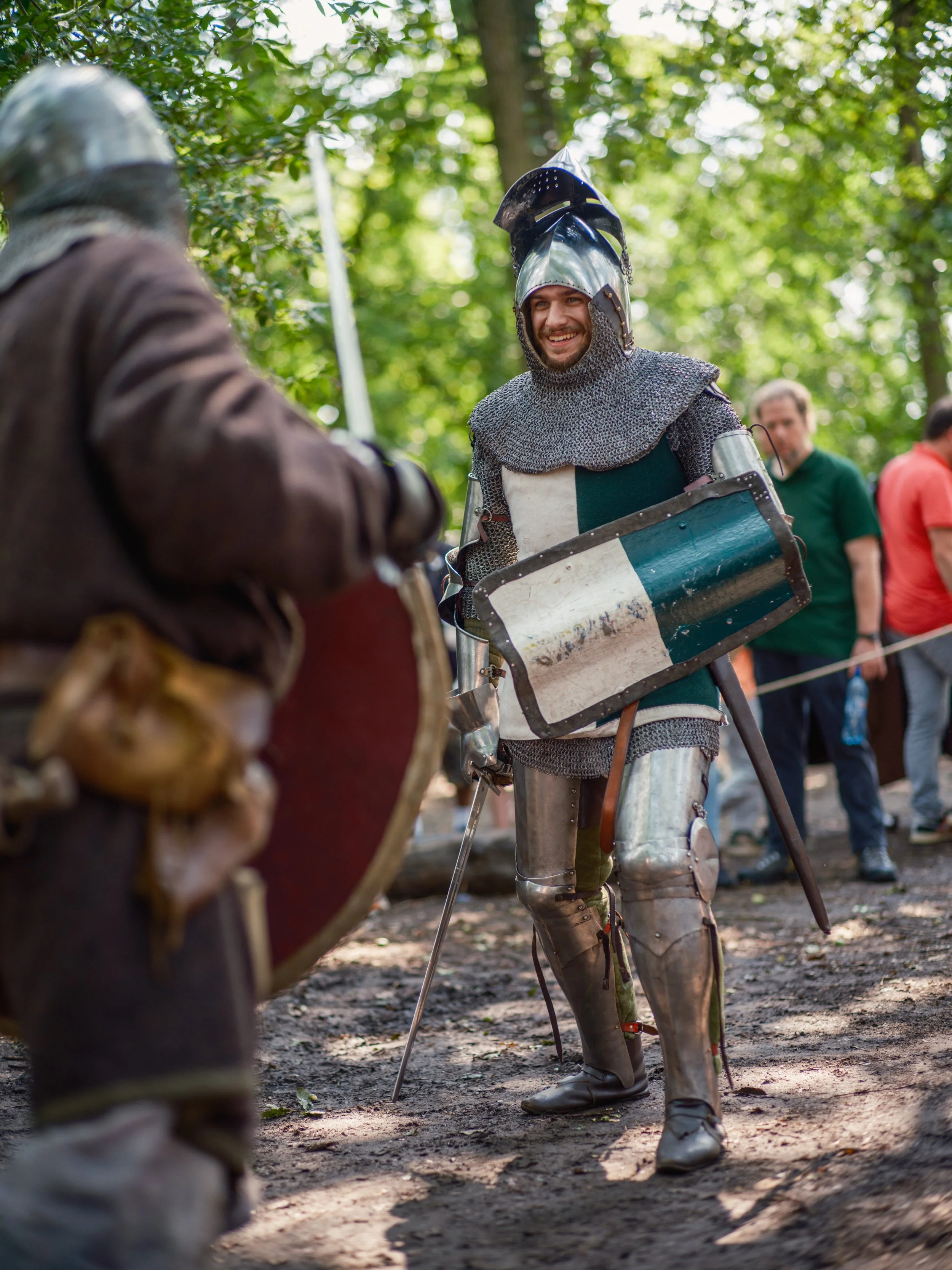 A smiling man dressed as a medieval knight, wearing armor and chainmail, holding a shield, and engaging in a reenactment or event in a forest setting during daytime.