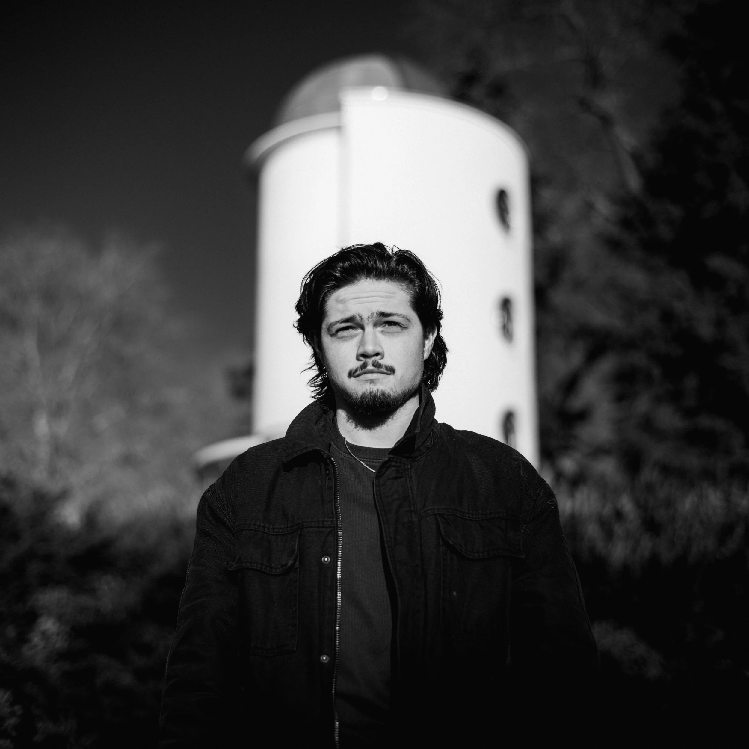 A black-and-white photo of a young man with wavy hair and a goatee, wearing a dark hooded jacket, standing outdoors with trees in the background.