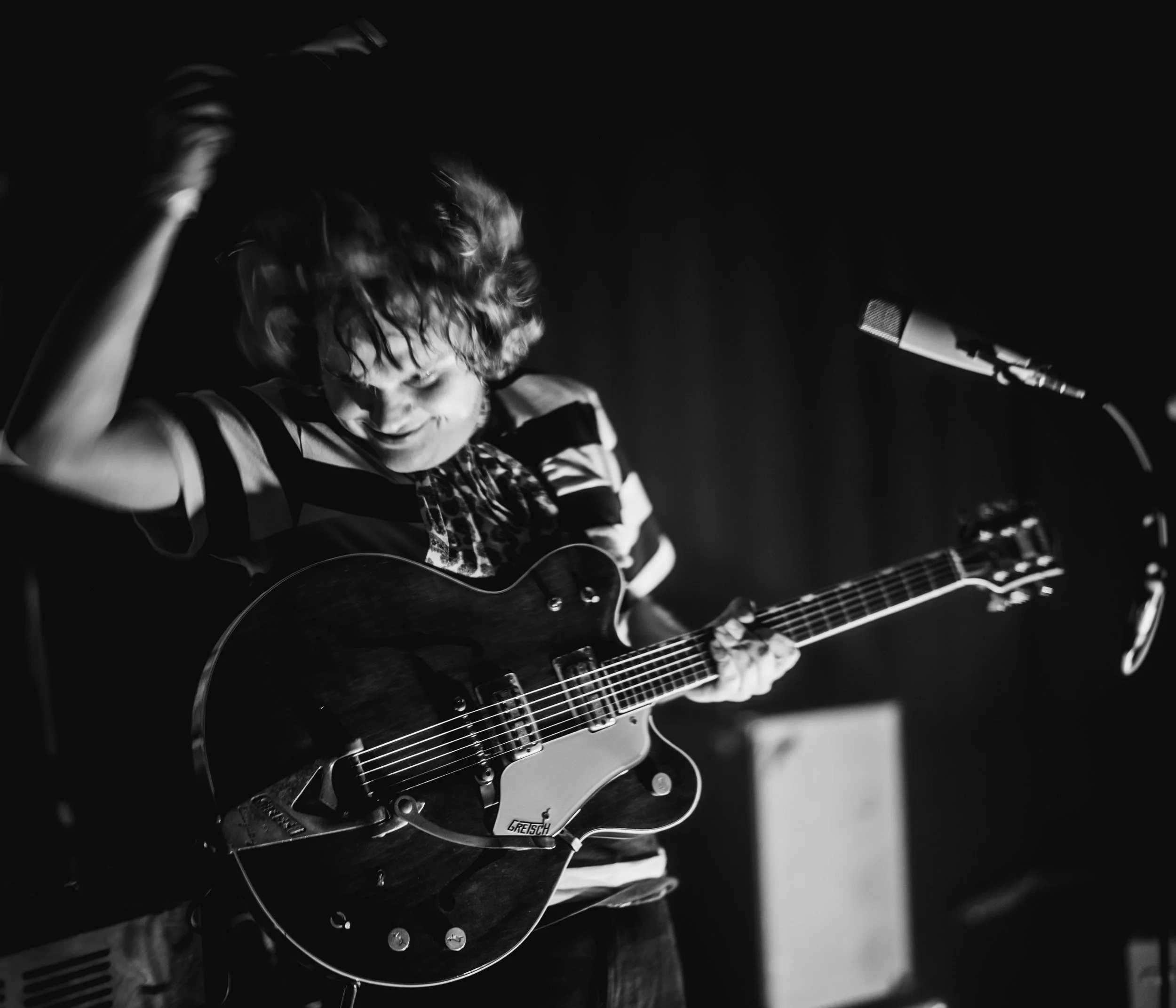 A person with curly hair wearing glasses, playing an electric guitar and holding a microphone in a dark setting.