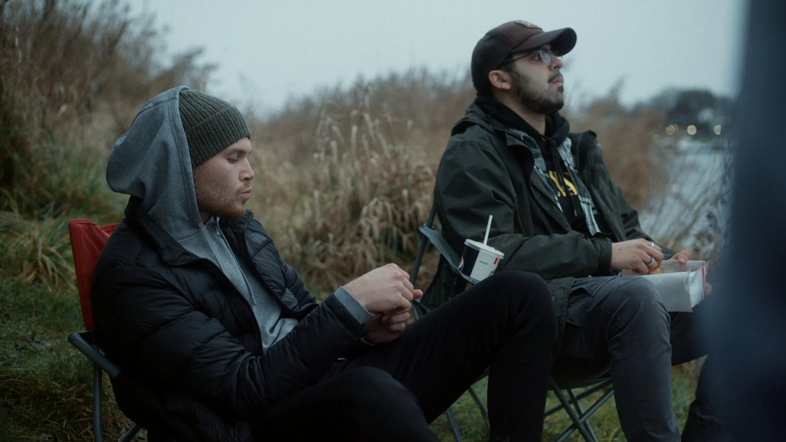 Two young men sitting outdoors on camping chairs near a body of water with tall grass behind them, one wearing a beanie and looking at his phone, the other wearing a cap and glasses, holding a drink and a bag of food.