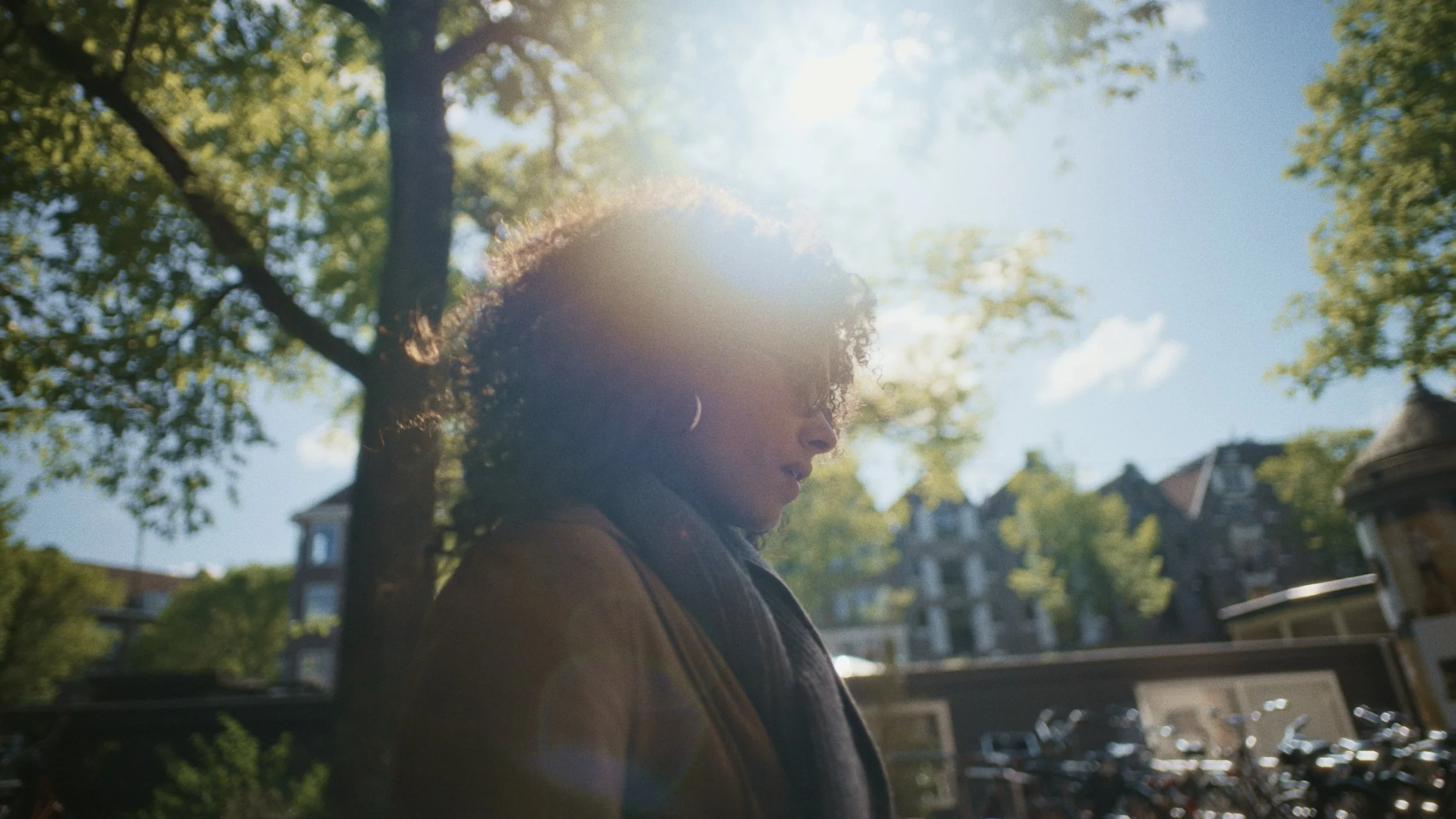 A woman with curly hair stands outdoors in sunlight, with trees and houses in the background.