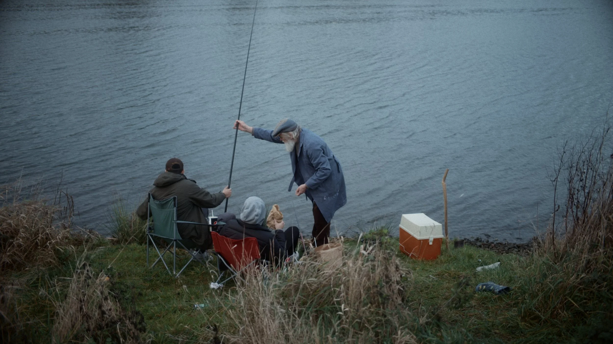 People fishing by a riverbank, some sitting in chairs, one woman standing, with fishing gear and a cooler nearby.