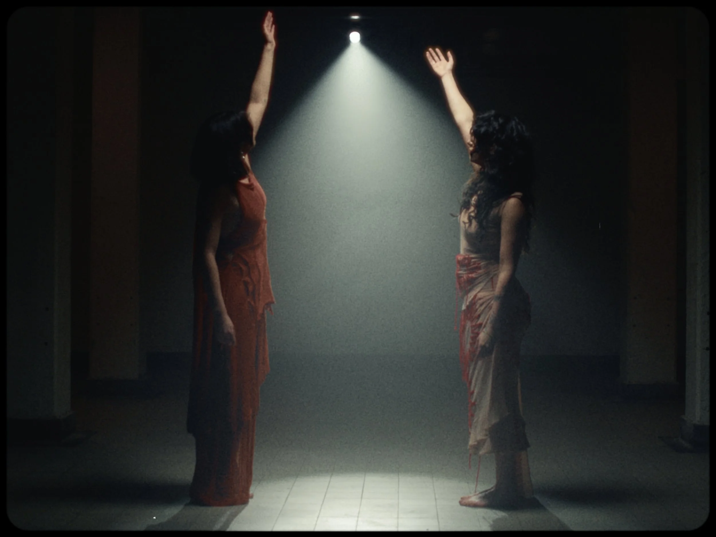Two women with curly hair are standing in a dark room, facing each other, with hand movements towards each other. They are illuminated by a spotlight from above. One woman wears an orange traditional outfit, and the other a patterned traditional skir