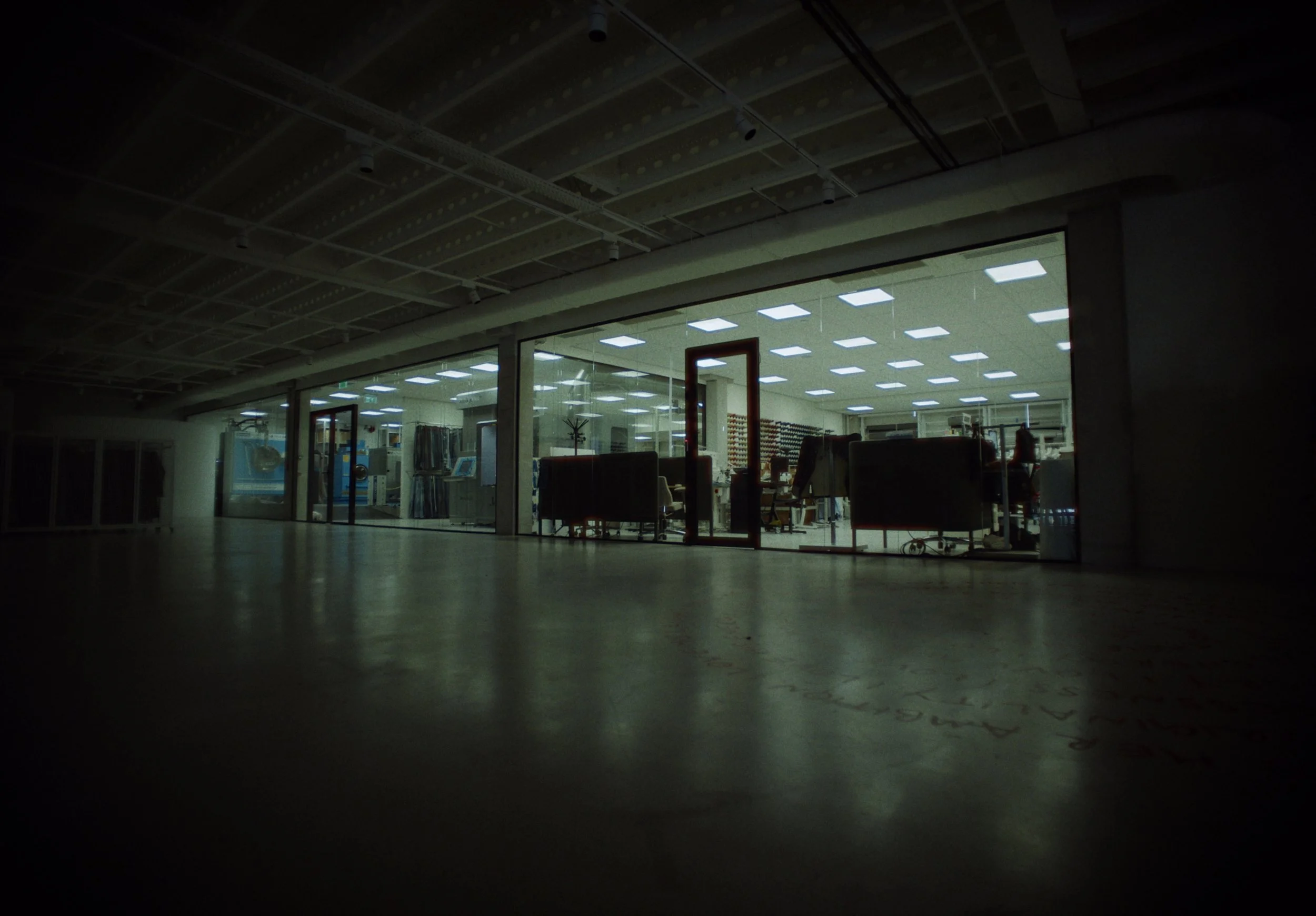 Dimly lit room with glass windows revealing a brightly lit interior space with ceiling lights, furniture, and equipment.