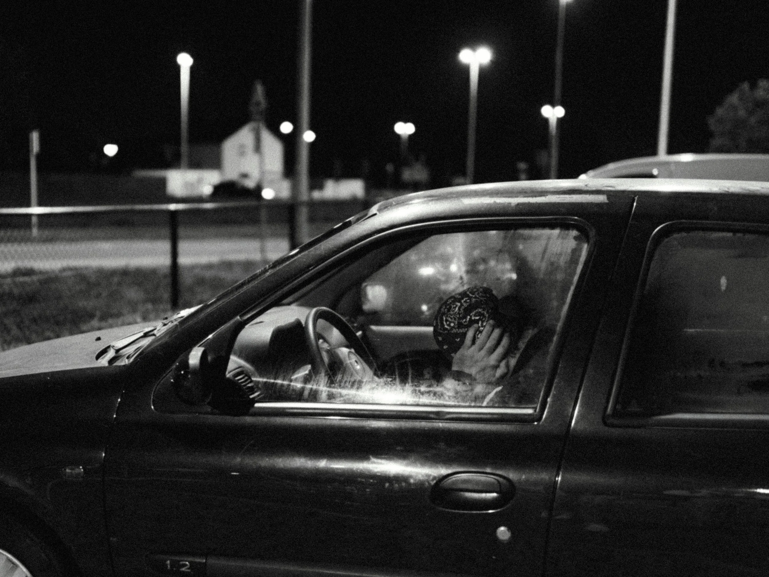 A person sitting inside a car at night, covering their face with a hand. The car is parked outdoors near streetlights and a small building, with a dark background.