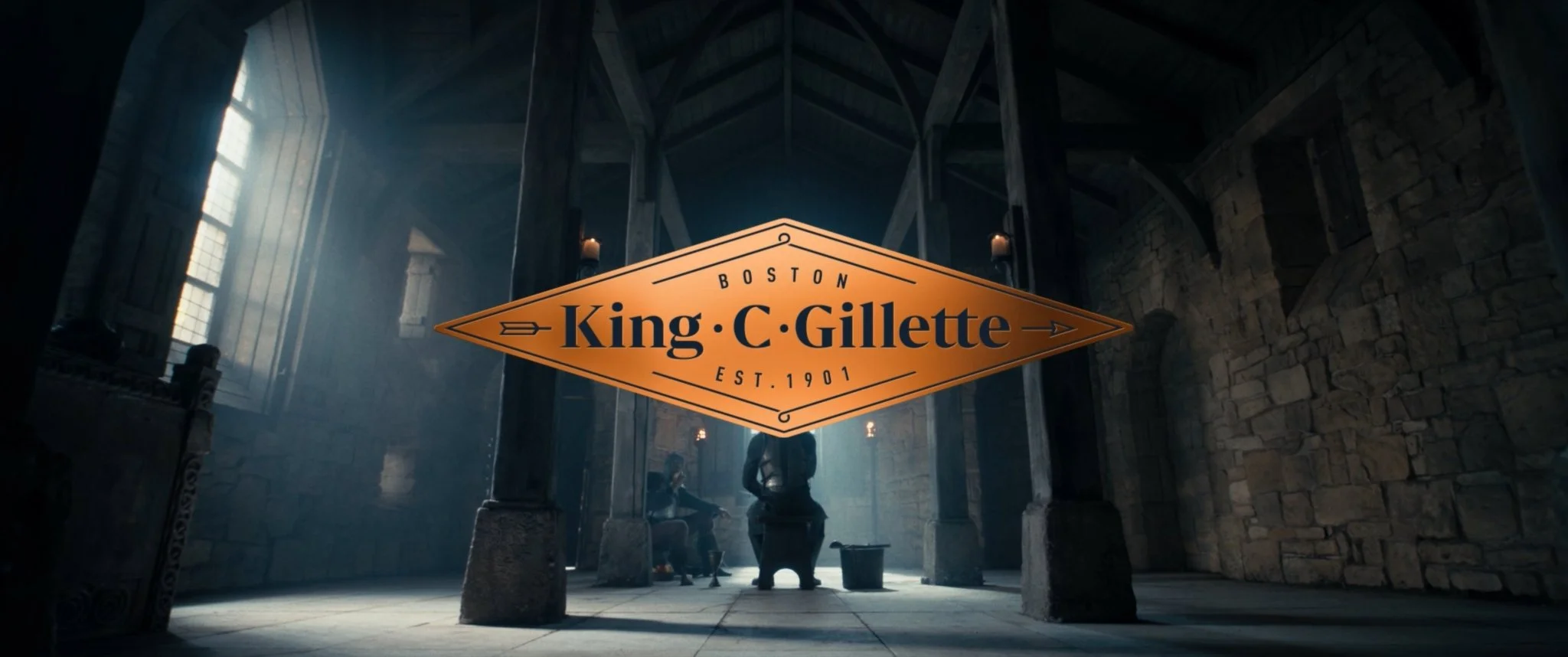 A large medieval-style hall with high wooden beams and stone walls. There are two candles on the wall. Two medieval figures sit near a fireplace. In the foreground, there is a prominent sign with the text 'Boston King C. Gillette Est. 1901'.