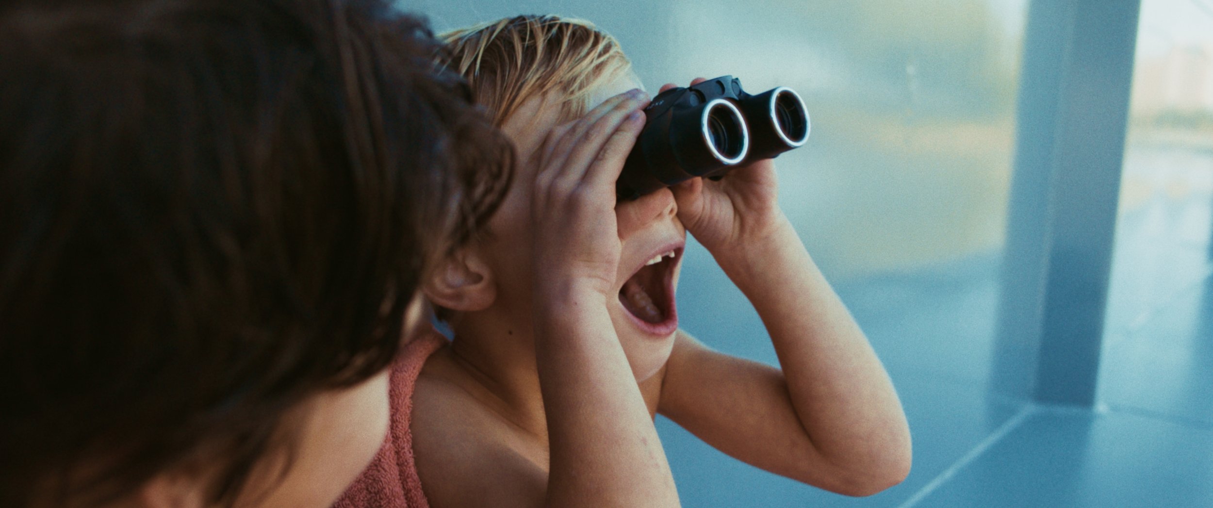 A young girl with blonde hair looking through binoculars with her mouth open, possibly excited or surprised, while another person with dark hair observes.