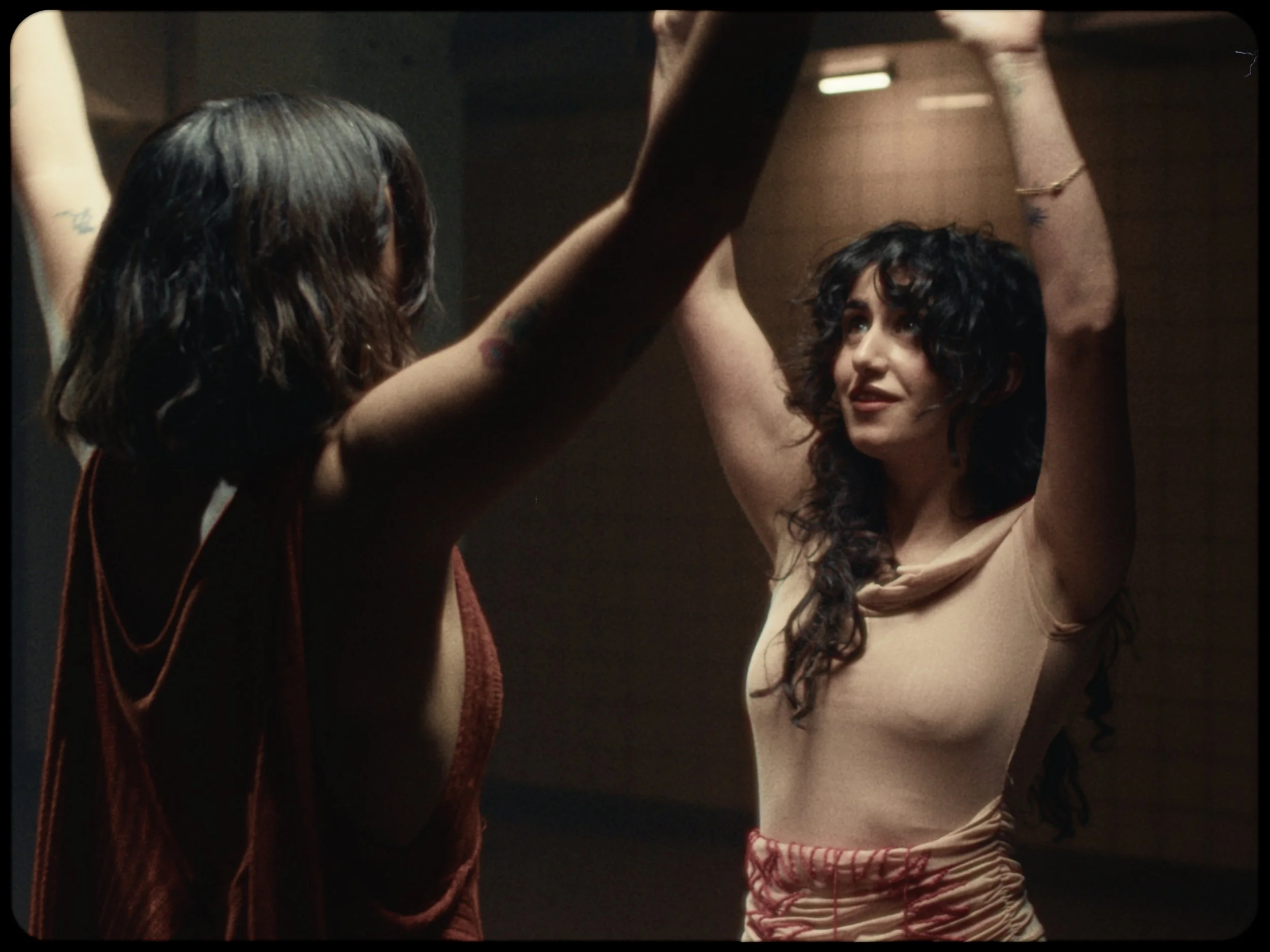 Two women, one with dark curly hair wearing a beige top and red-striped shorts, and the other with long brown hair wearing a rust-colored sleeveless top, are dancing together with their arms raised.
