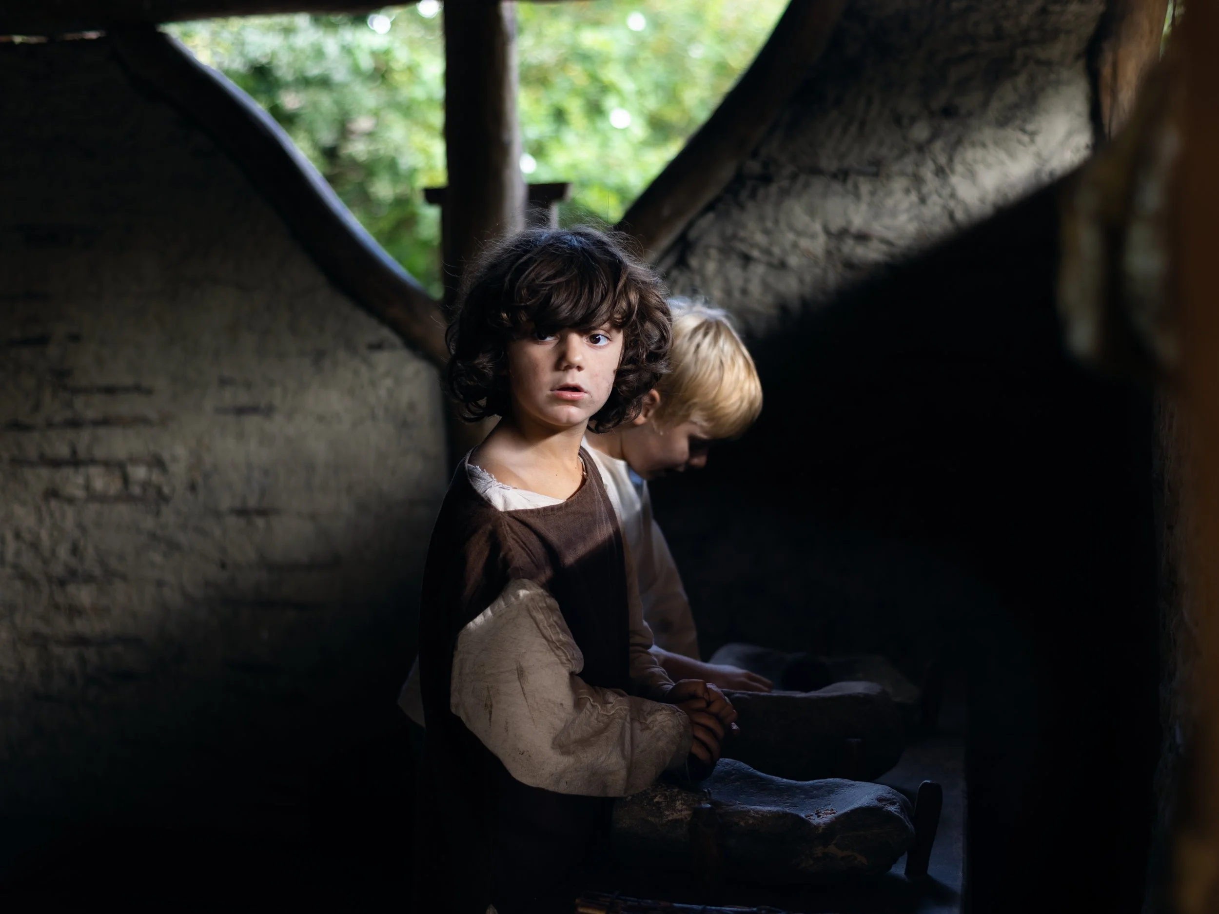 Two children, a girl with dark curly hair and a boy with light hair, sit inside a dimly lit wooden structure, possibly a hut or shelter, with greenery visible outside.