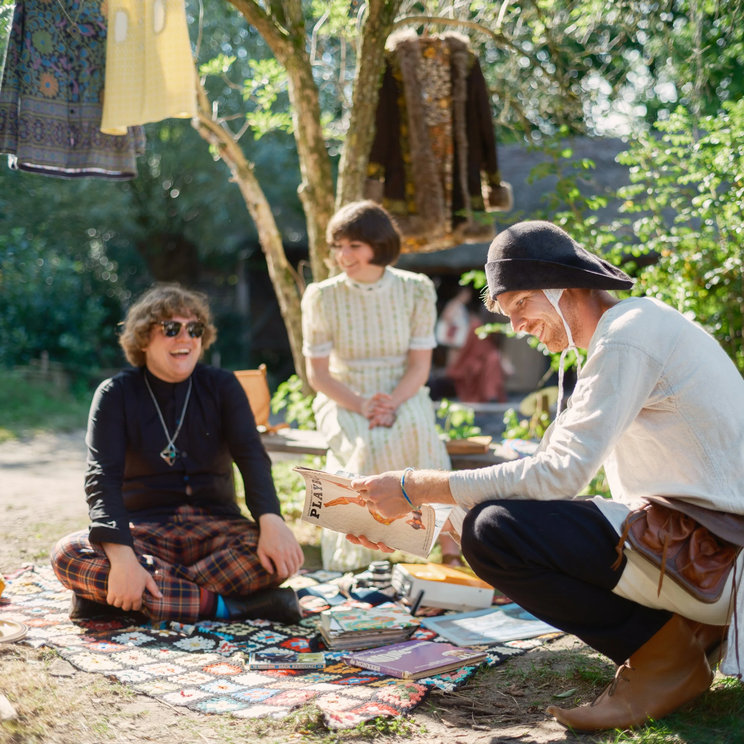 People sitting outdoors on a colorful rug, reading magazines and comics, with a background of trees and sunlight filtering through leaves.