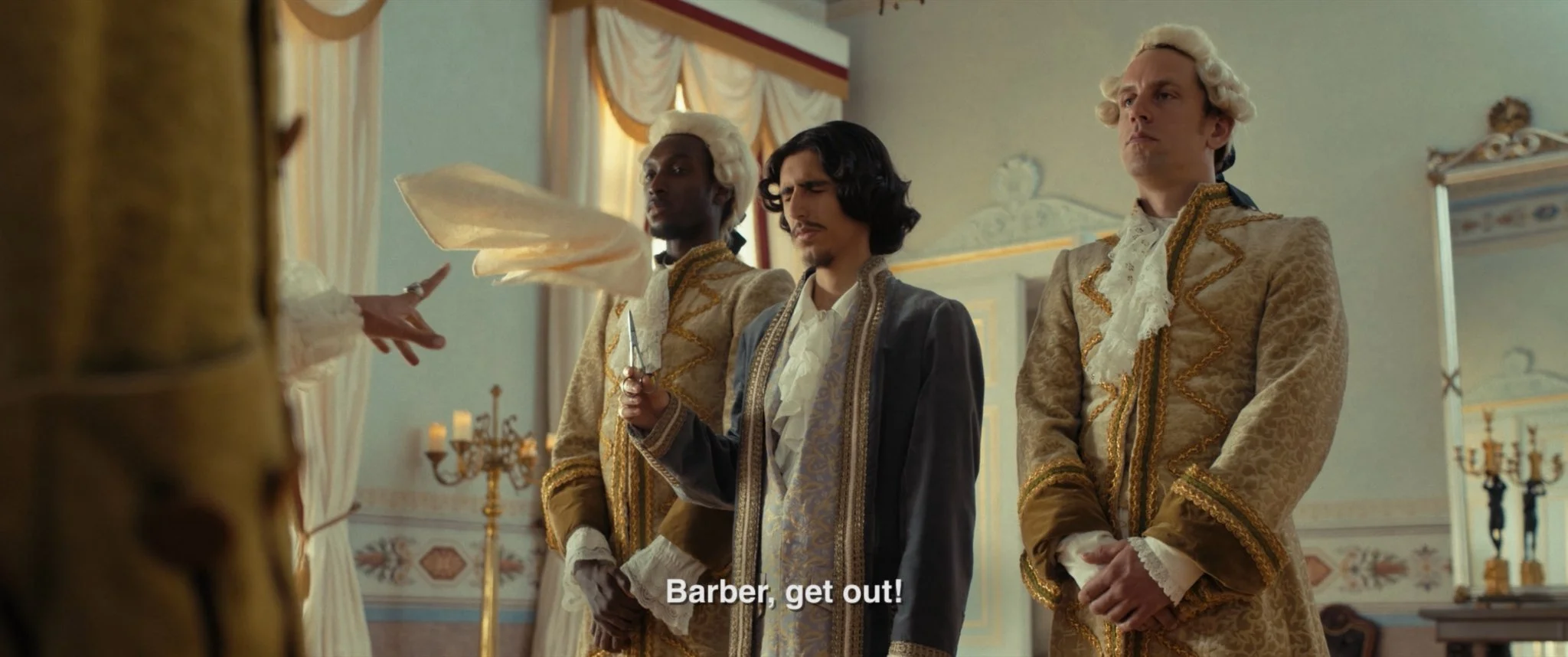 Three men dressed in elaborate 18th-century aristocratic clothing standing in a grand, decorated room with chandeliers and draped curtains. One man in the center appears upset, holding a dagger, while the other two stand with serious expressions. A w