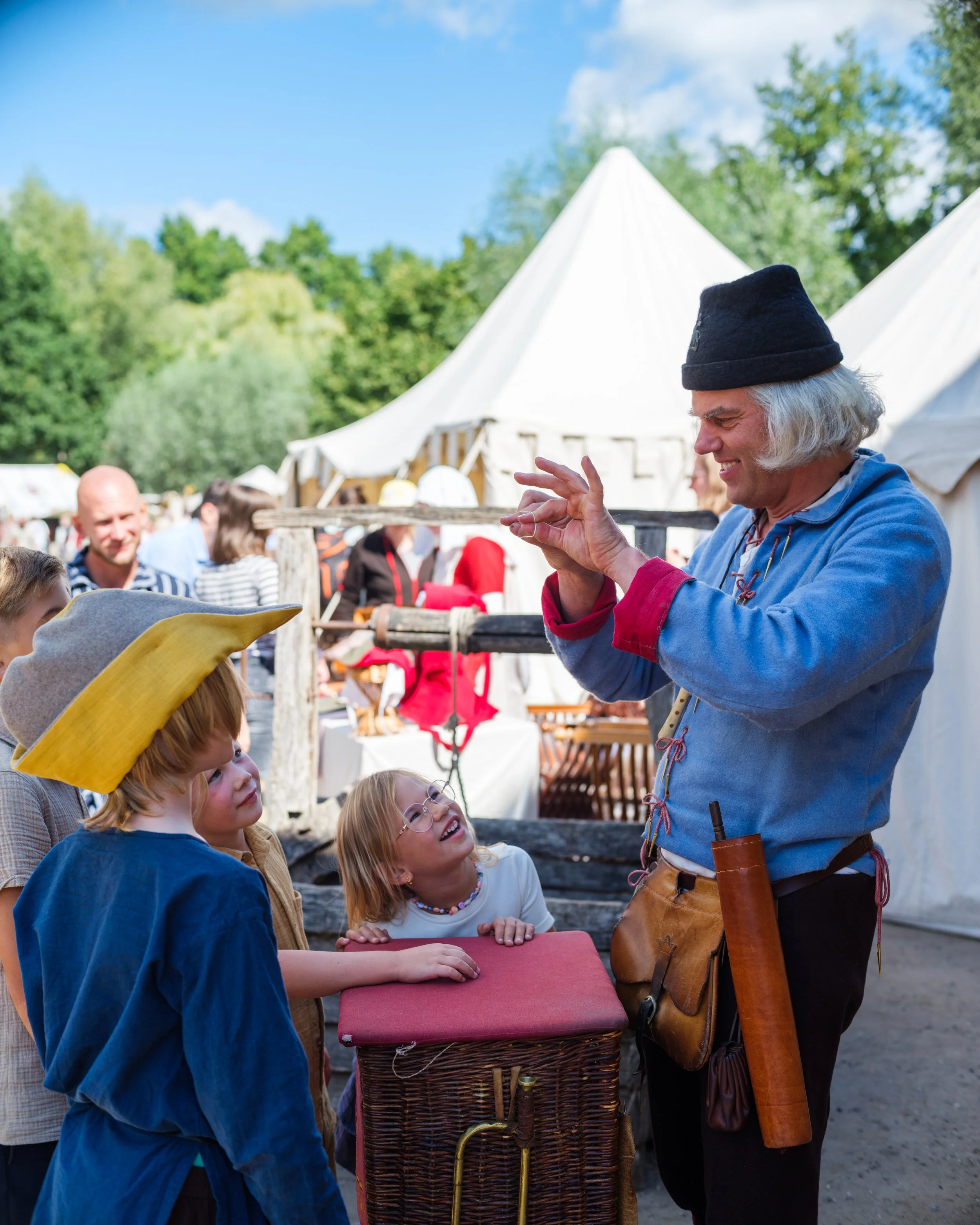 An elderly man in a medieval costume with a black hat and a blue shirt, smiling and showing something to a group of children at an outdoor event, with white tents and green trees in the background.
