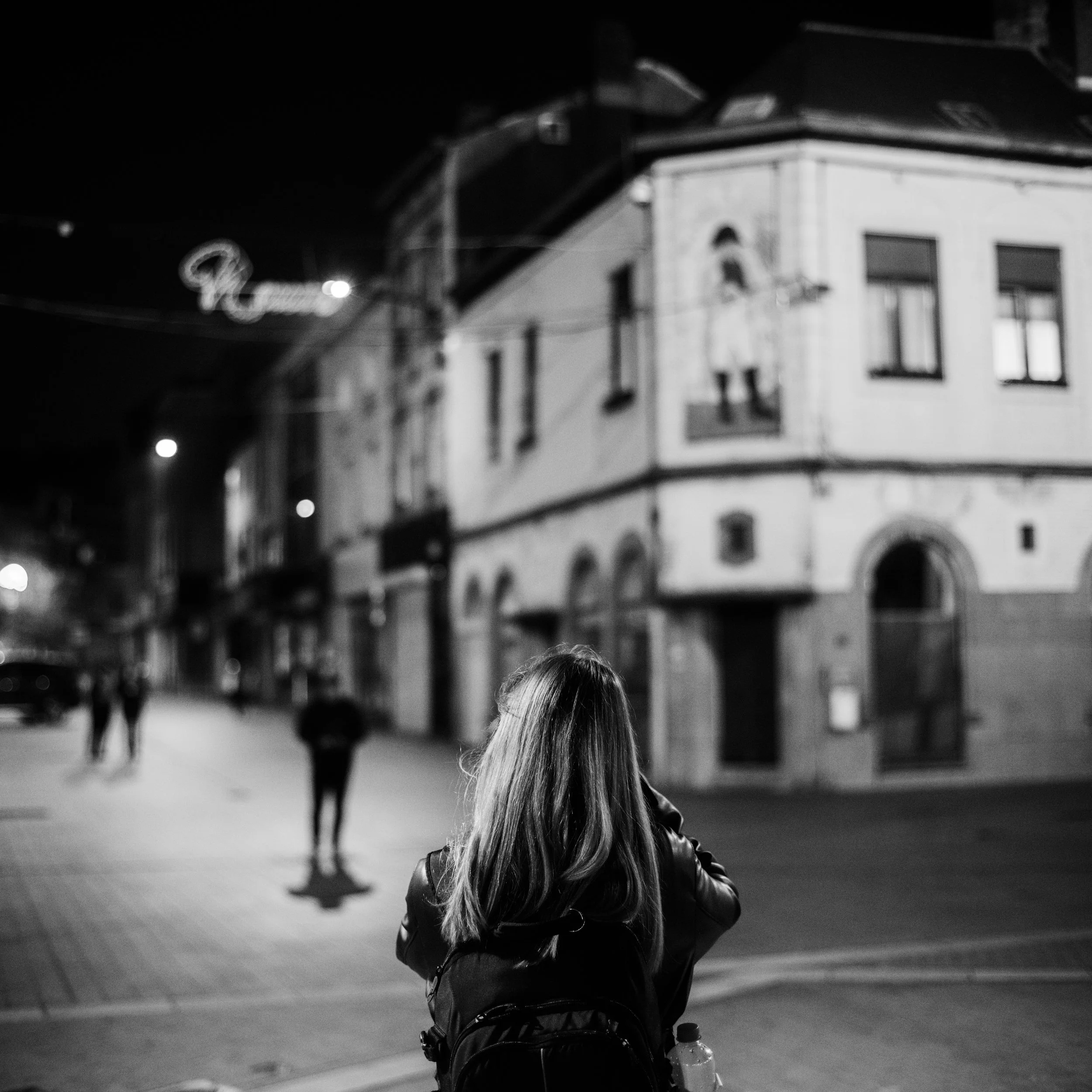 A girl with long hair wearing a jacket standing on a city street at night, facing away from the camera, with a building and illuminated signs in the background.