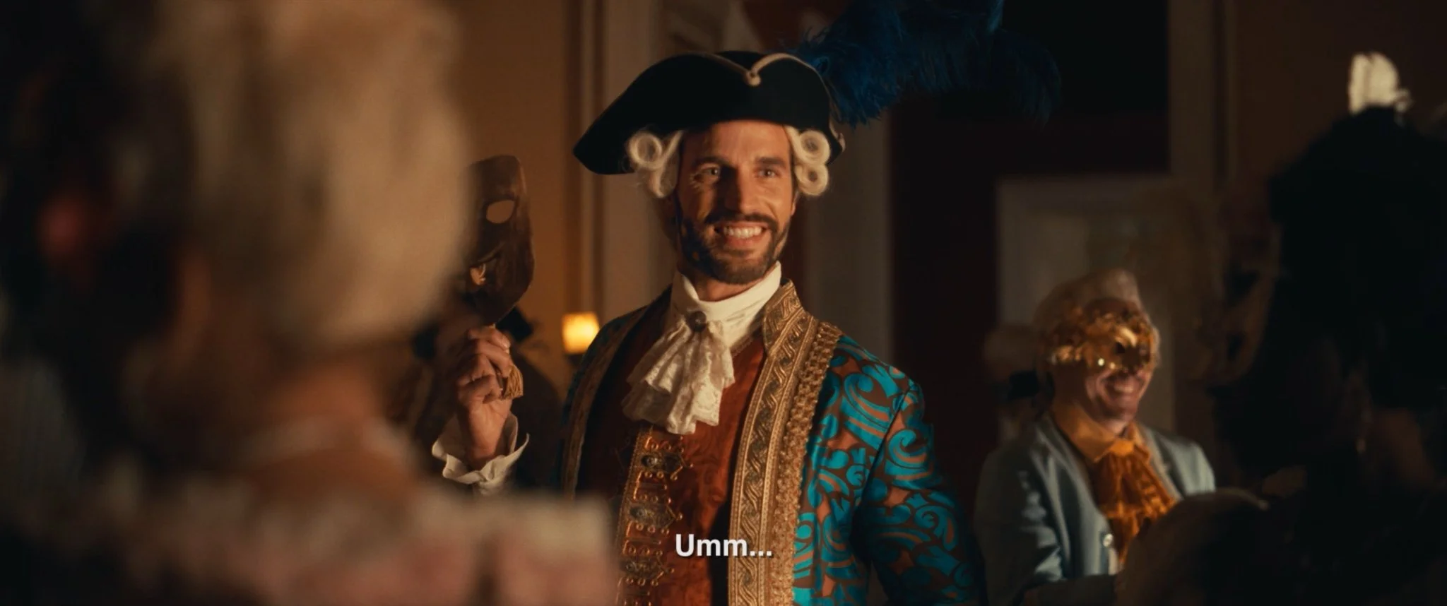 A man in 18th-century aristocratic attire, smiling in a social gathering, wearing a large powdered wig, a tricorn hat with a feather, and a decorative blue and gold coat.