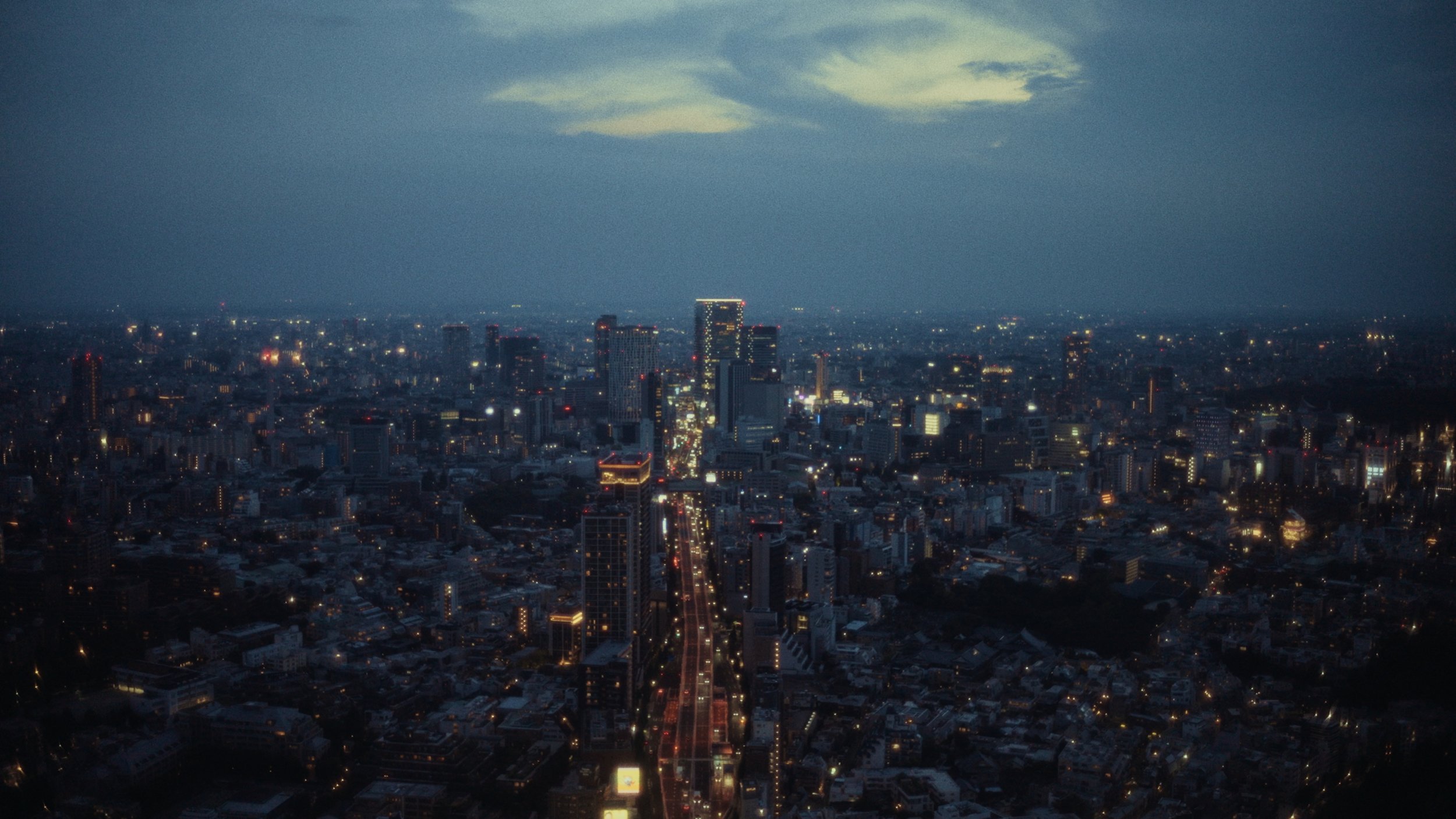Aerial view of a city at night with illuminated buildings and streets, and a cloudy sky overhead.