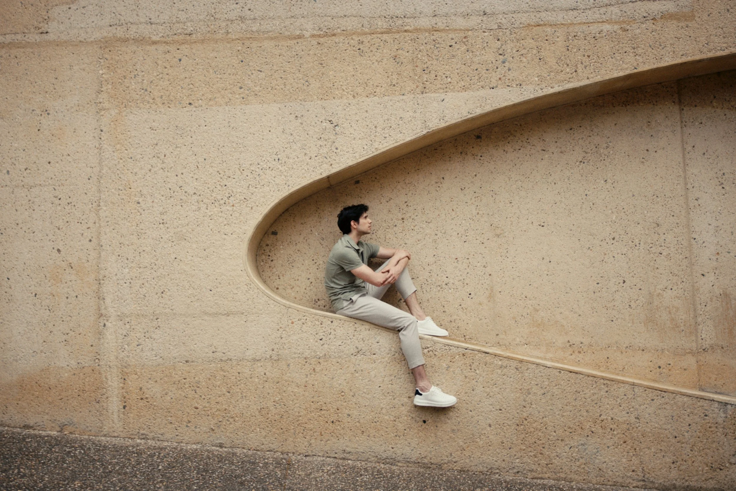 A young man sitting on a ledge with a large curved opening in a beige textured wall, wearing a casual gray outfit and white sneakers.