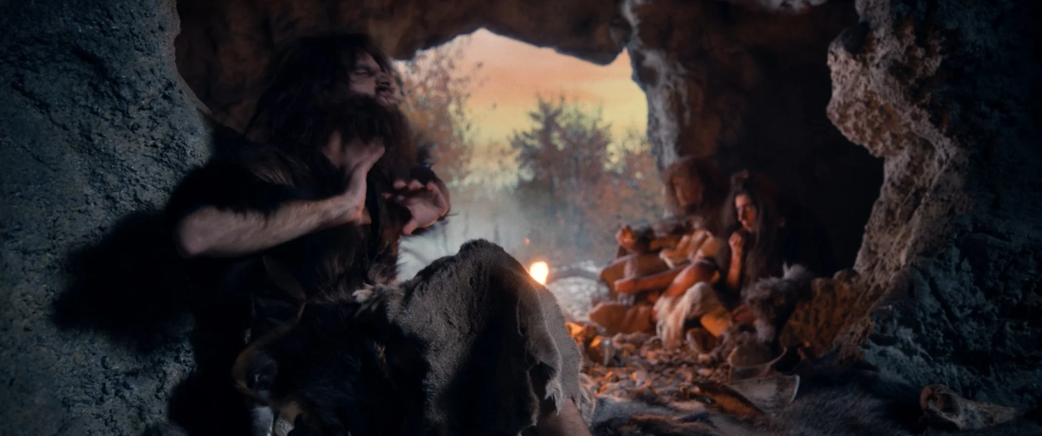 Two characters, one with long hair and a beard and one with shorter hair, are sitting in a rocky cave. The person with long hair appears to be upset or distressed, while the other is holding them. The scene is set during sunset, with sunlight filteri