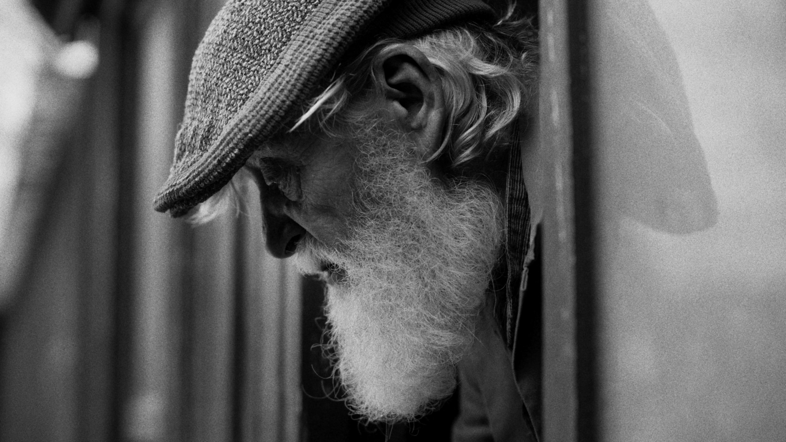 Close-up black and white photo of an elderly man with a long white beard wearing a hat, with his head slightly bowed and his face in profile.