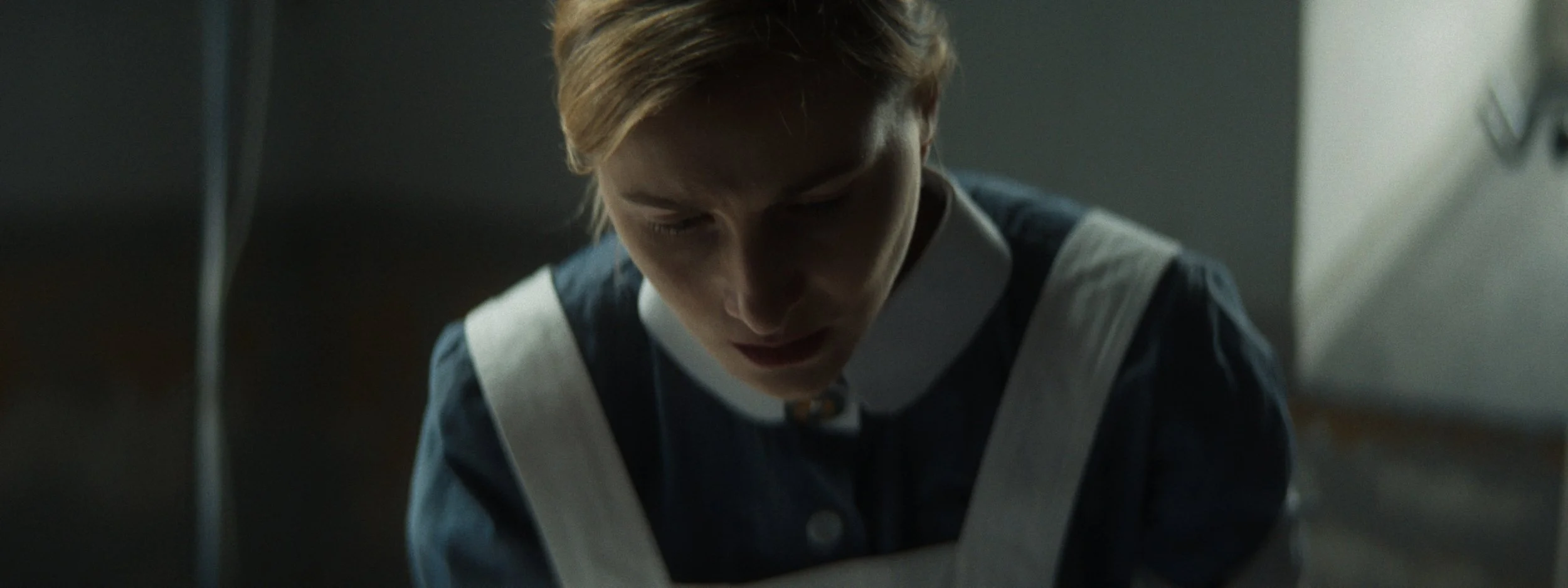 A woman with short blonde hair looking down, wearing a dark uniform with white straps, in a dimly lit room.