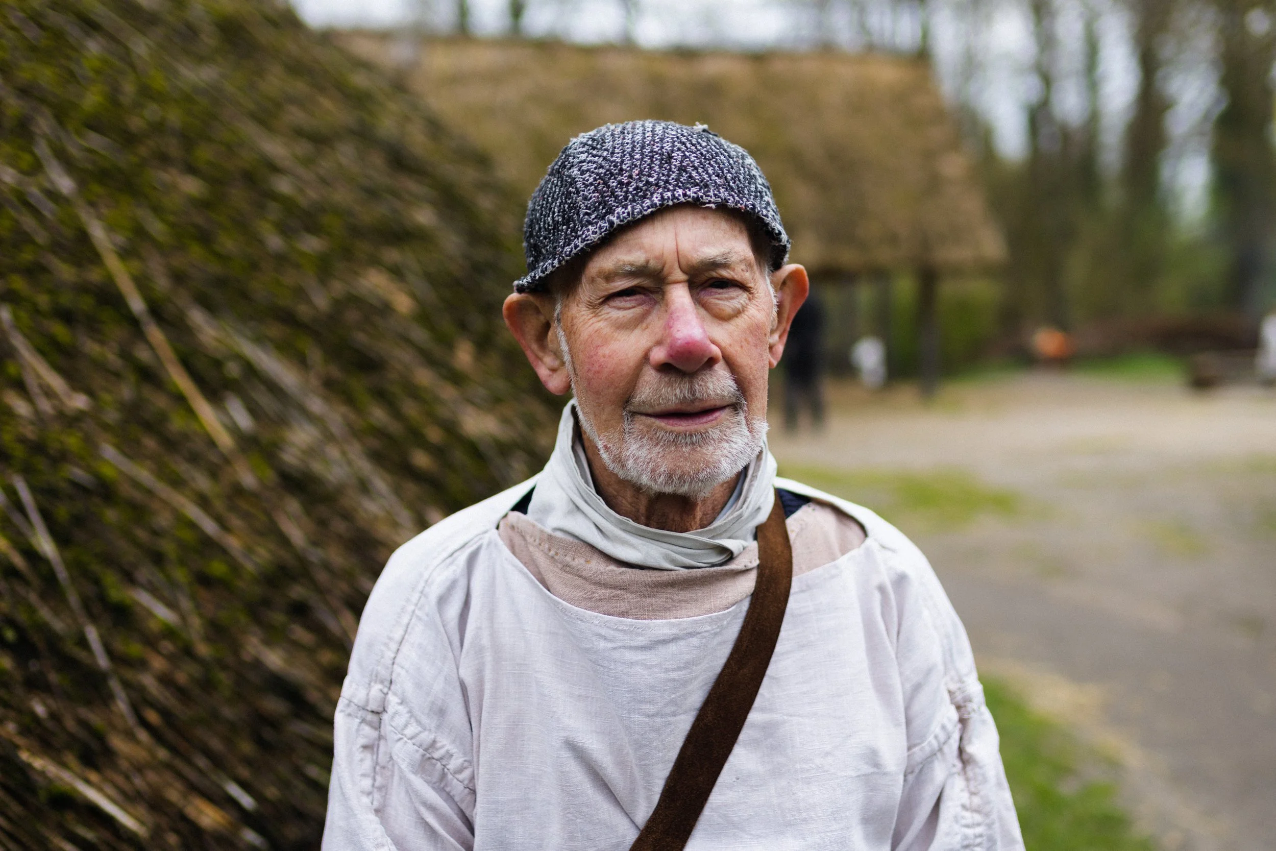 An elderly man with a gray beard wearing a gray knit cap and a light-colored shirt, outdoors in a park or garden with blurred trees and a building in the background.