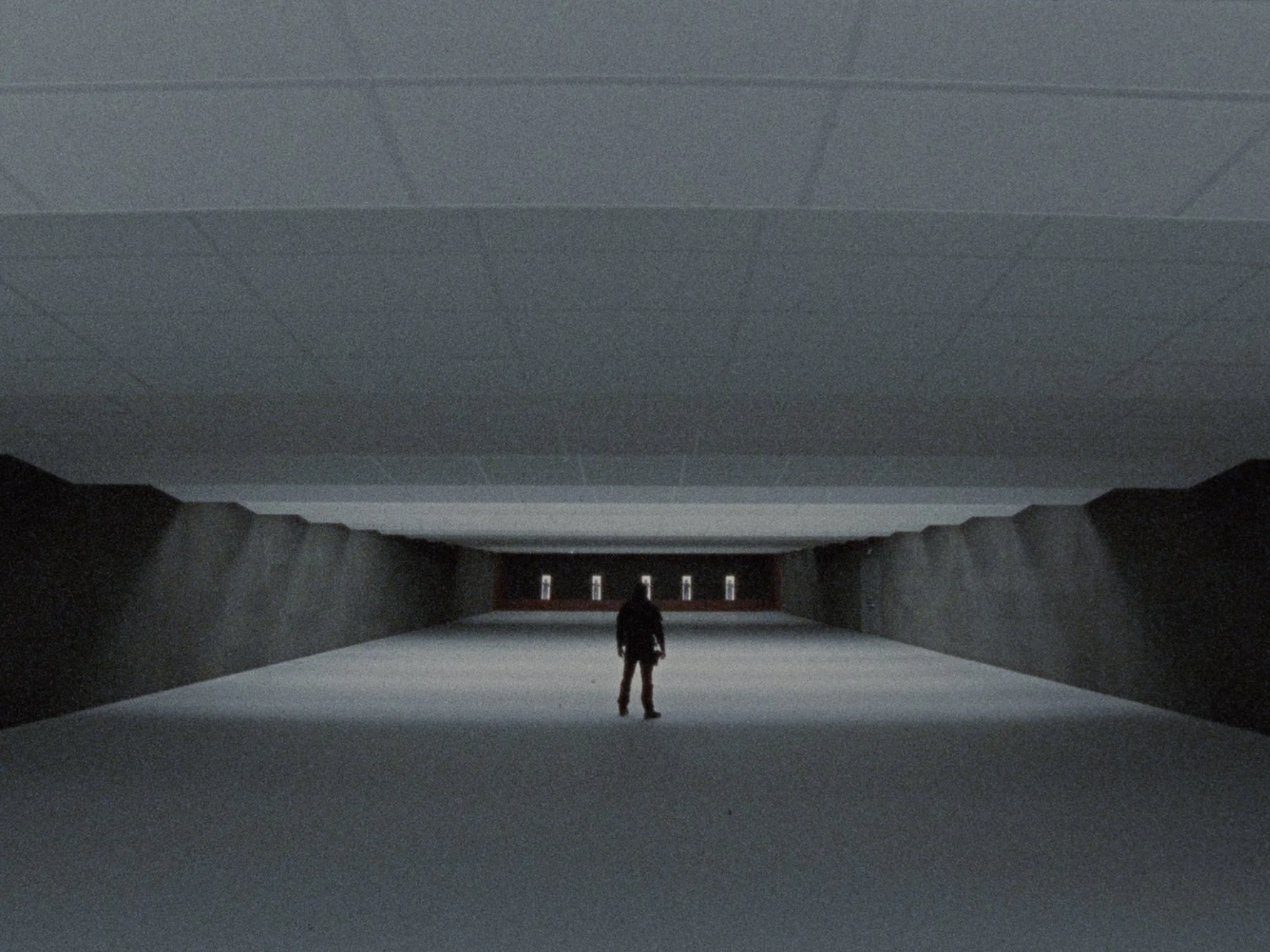 A person standing in a dark, empty tunnel or corridor with a high ceiling and minimal lighting, looking towards the opening at the end of the tunnel.