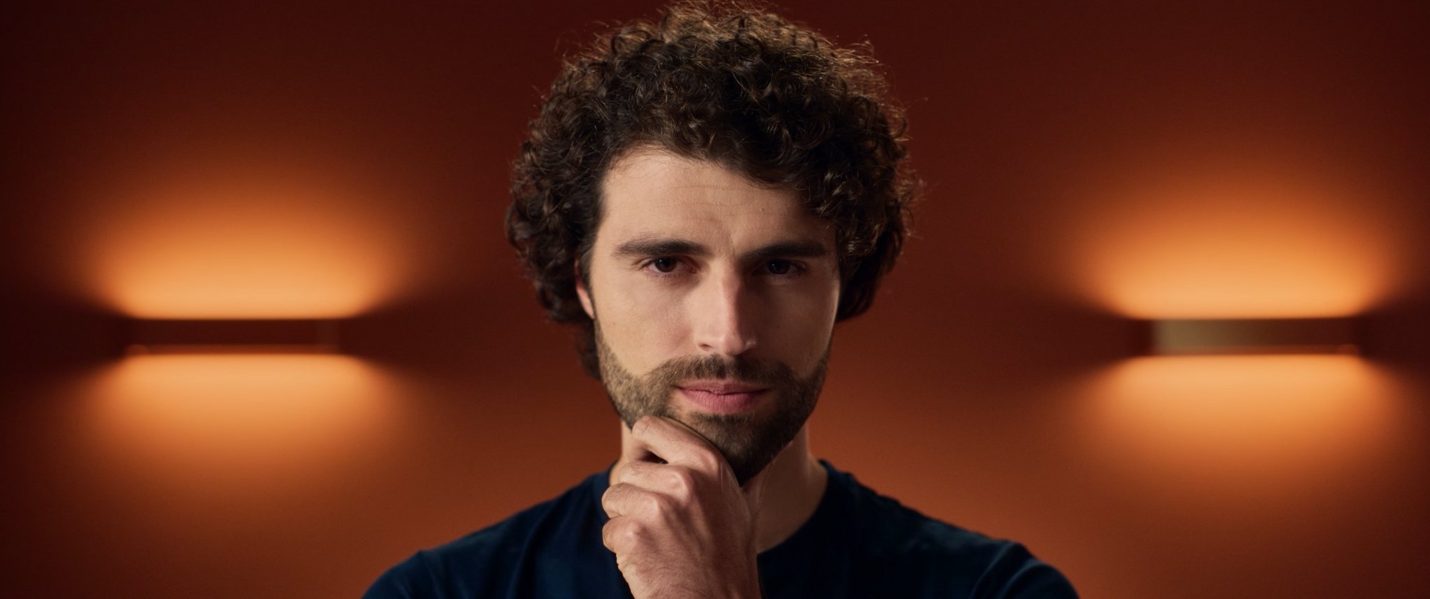 A man with curly hair and a beard, resting his chin on his hand, looking directly at the camera against a blurred warm-toned background.