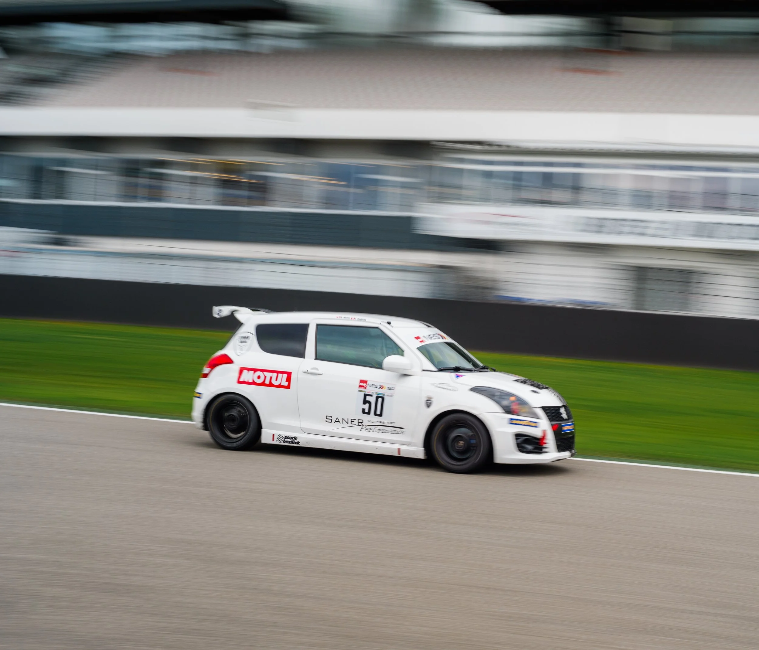 A white racing car with sponsorship logos speeding on a race track, with blurred background showing infrastructure and other vehicles.