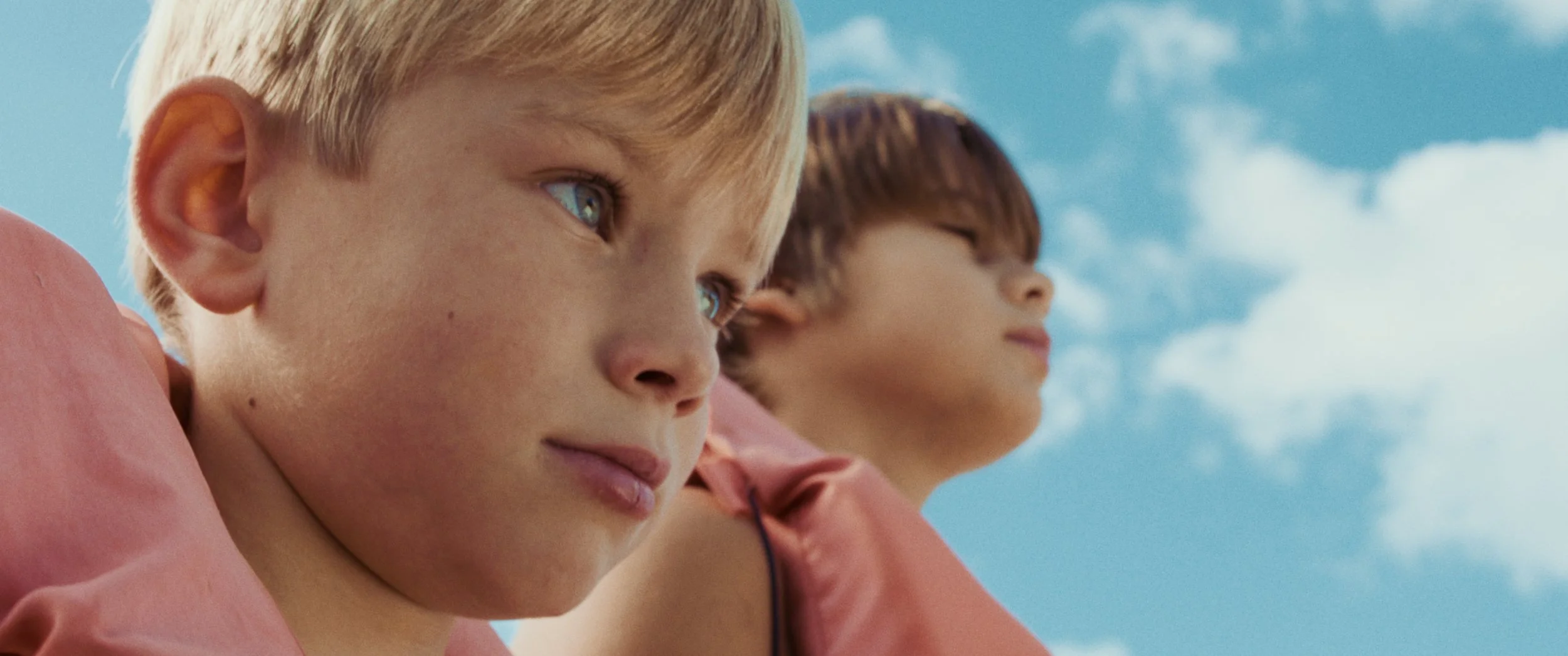 Close-up of two young boys wearing pink shirts looking off into the distance against a blue sky with clouds.