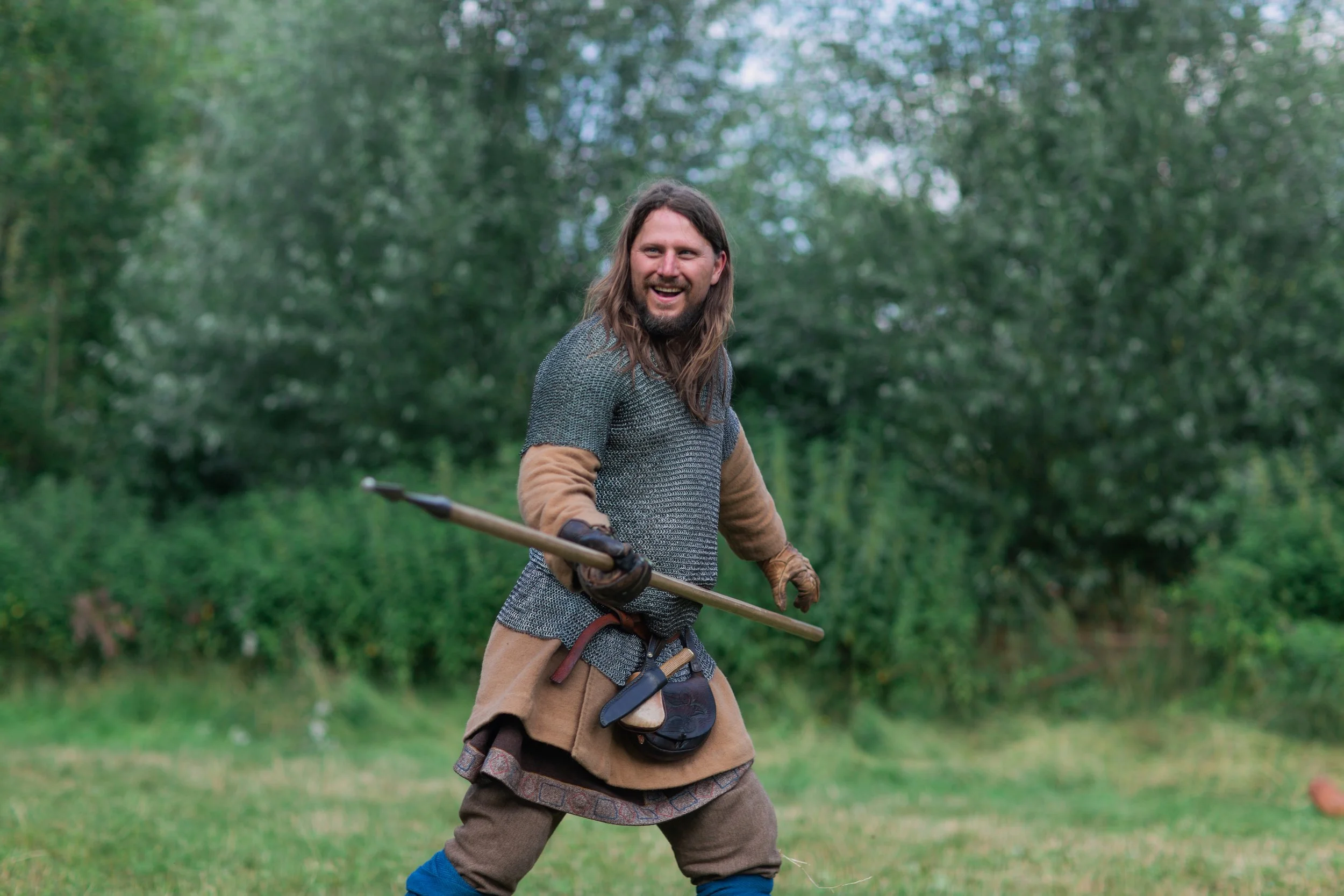 A man with long hair and a beard dressed in medieval style clothing holding a spear outdoors amid greenery, smiling.
