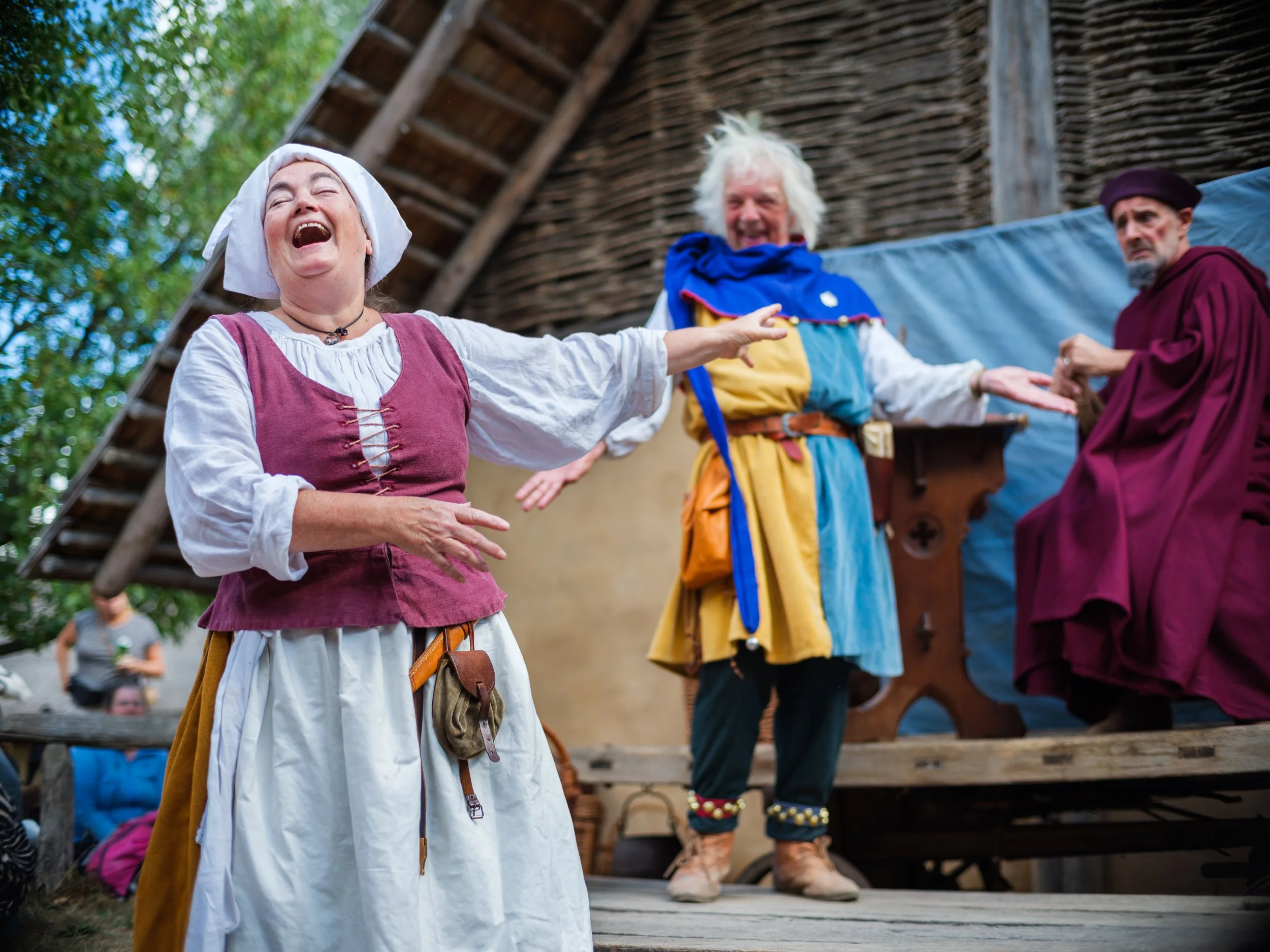 A woman dressed as a medieval character, laughing and pointing, with two men in medieval costumes behind her, on a stage with a thatched roof and wooden structure.