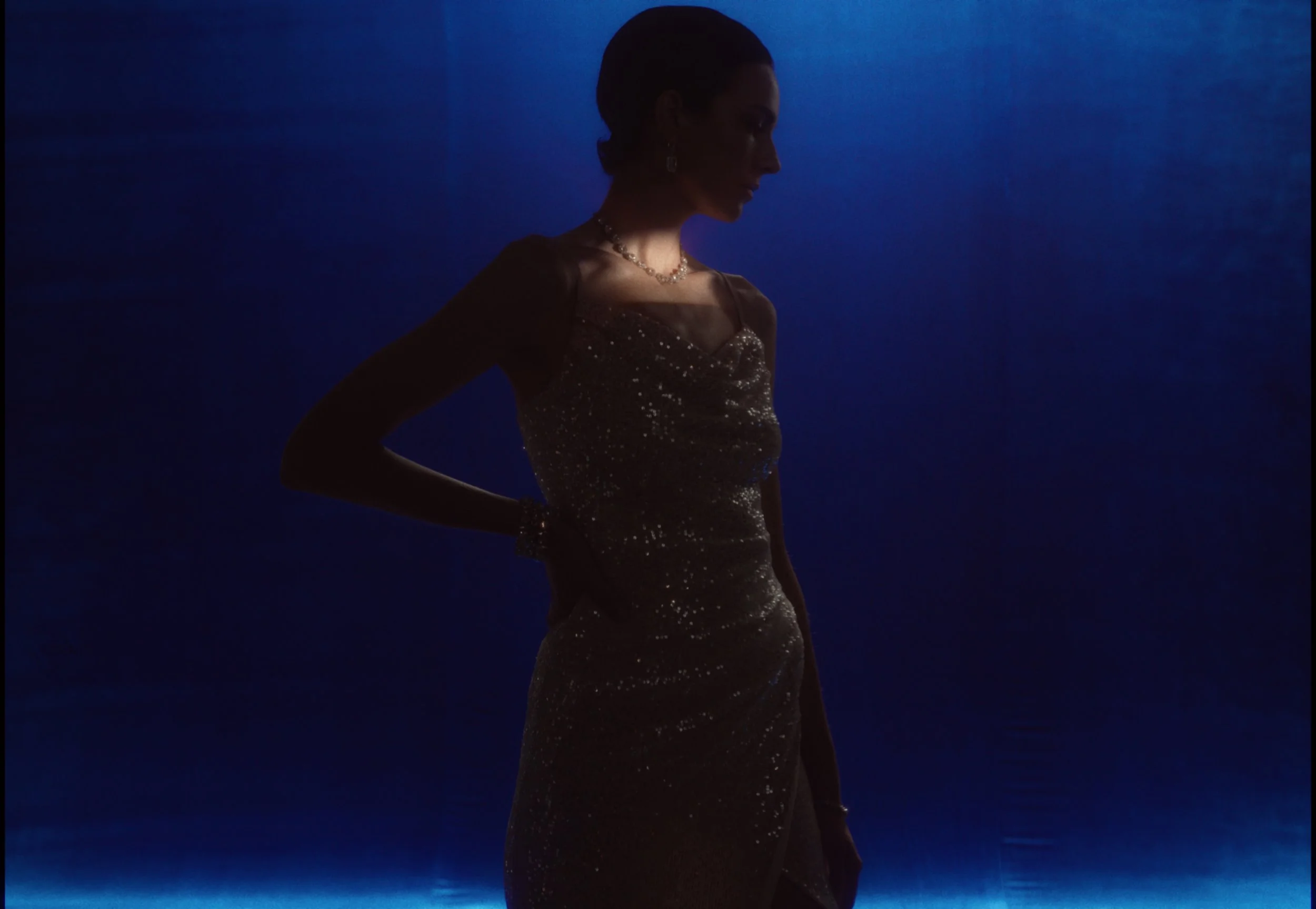 A woman dressed in a glamorous, sequined gown, standing in front of a dark blue background. She is accessorized with jewelry and appears to be in a contemplative pose with her head slightly bowed.