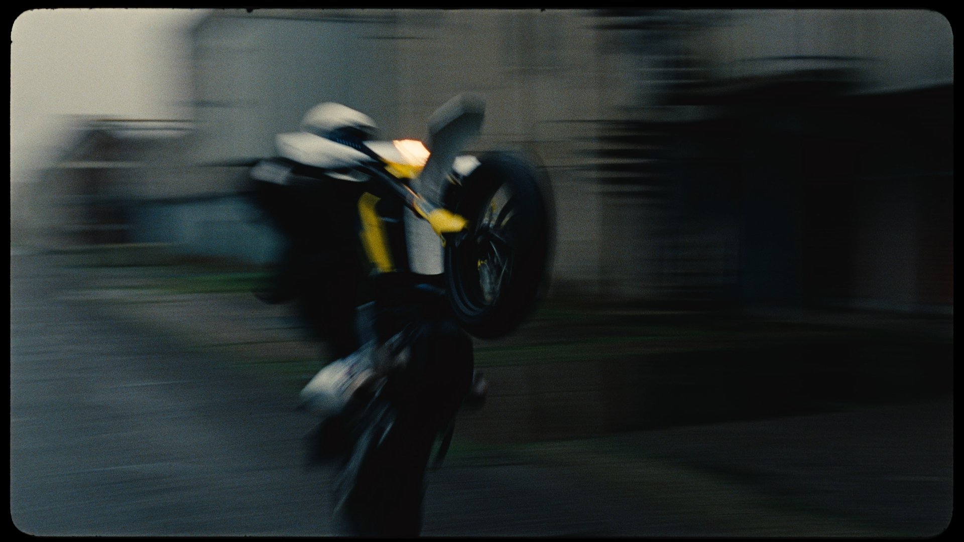 A person performing a stunt on a motorcycle, lifting the front wheel off the ground while balancing on the rear wheel inside a garage with shelves and equipment in the background