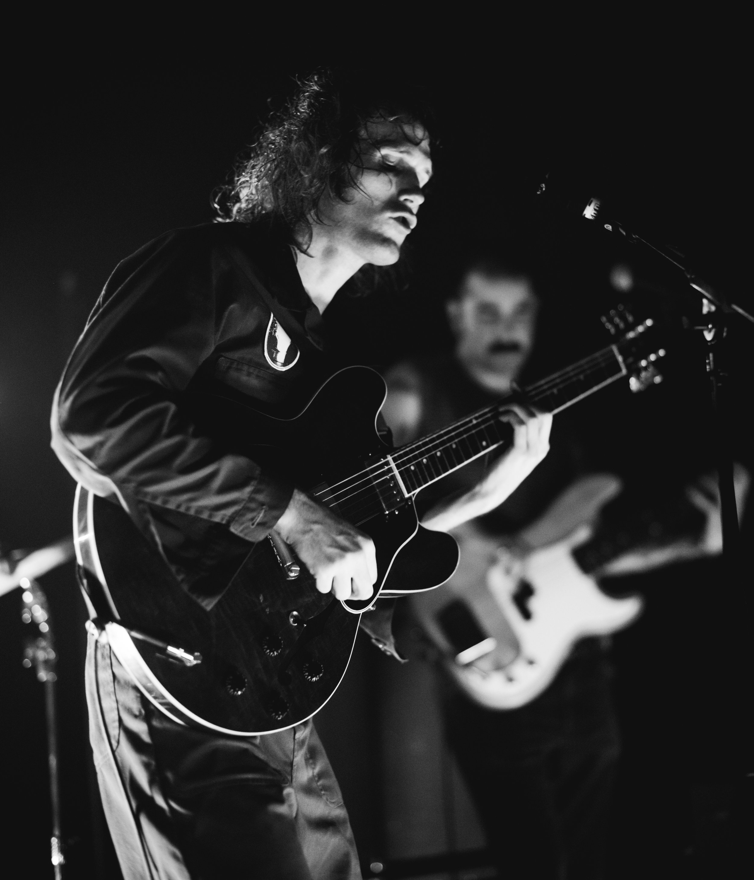 A man with curly hair playing an acoustic guitar on stage, with a blurred background, black and white photo.
