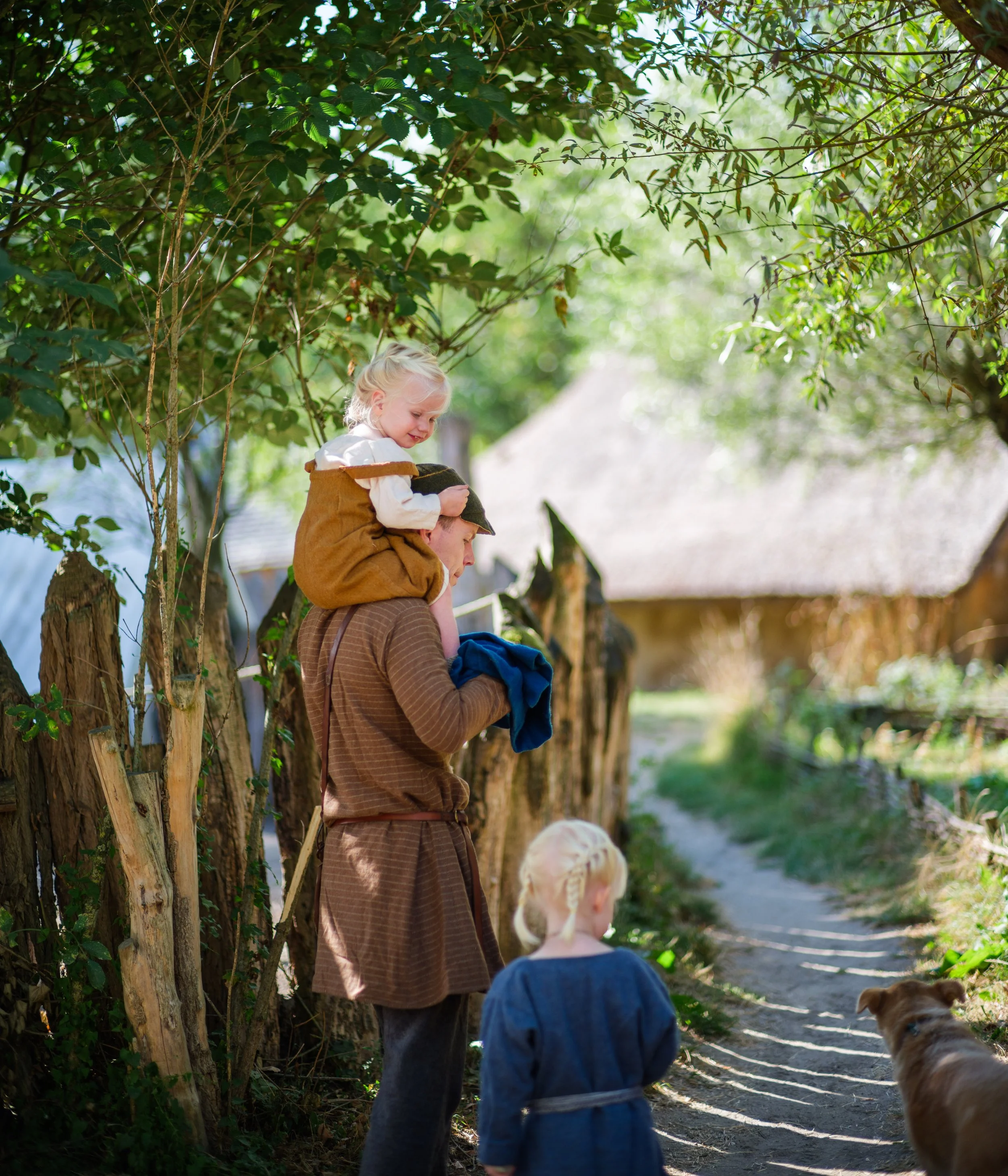 A young woman carrying a small blonde girl on her shoulders while walking along a shaded path with children and dogs nearby, surrounded by trees and greenery.