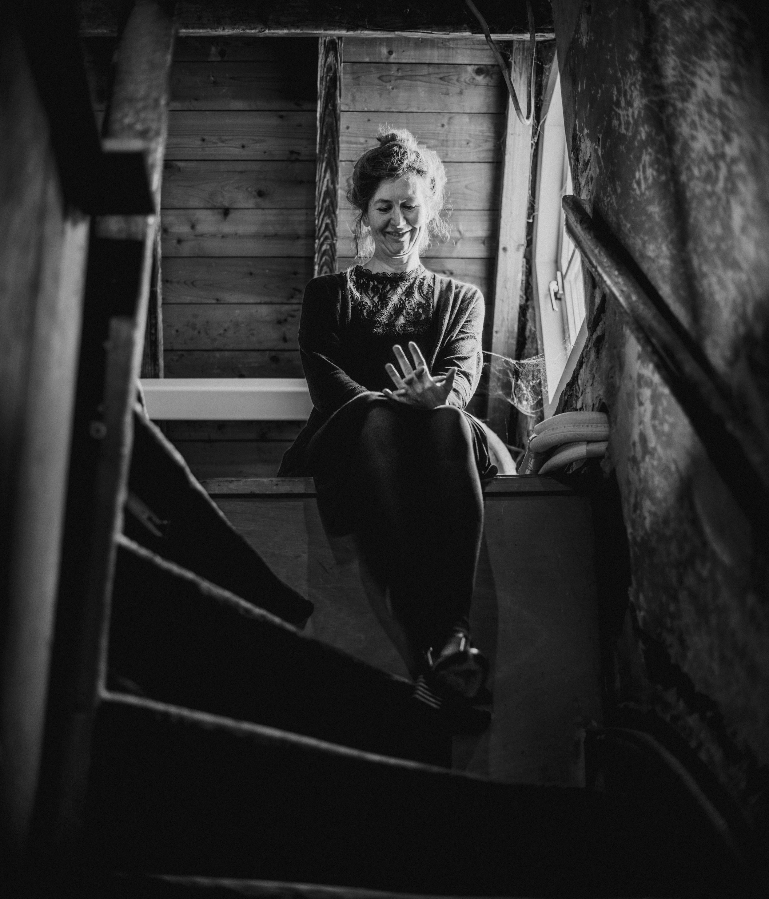 A woman with curly hair and a lace top, smiling and gesturing with her hand, sitting in a rustic room with wooden walls and a window to the side.
