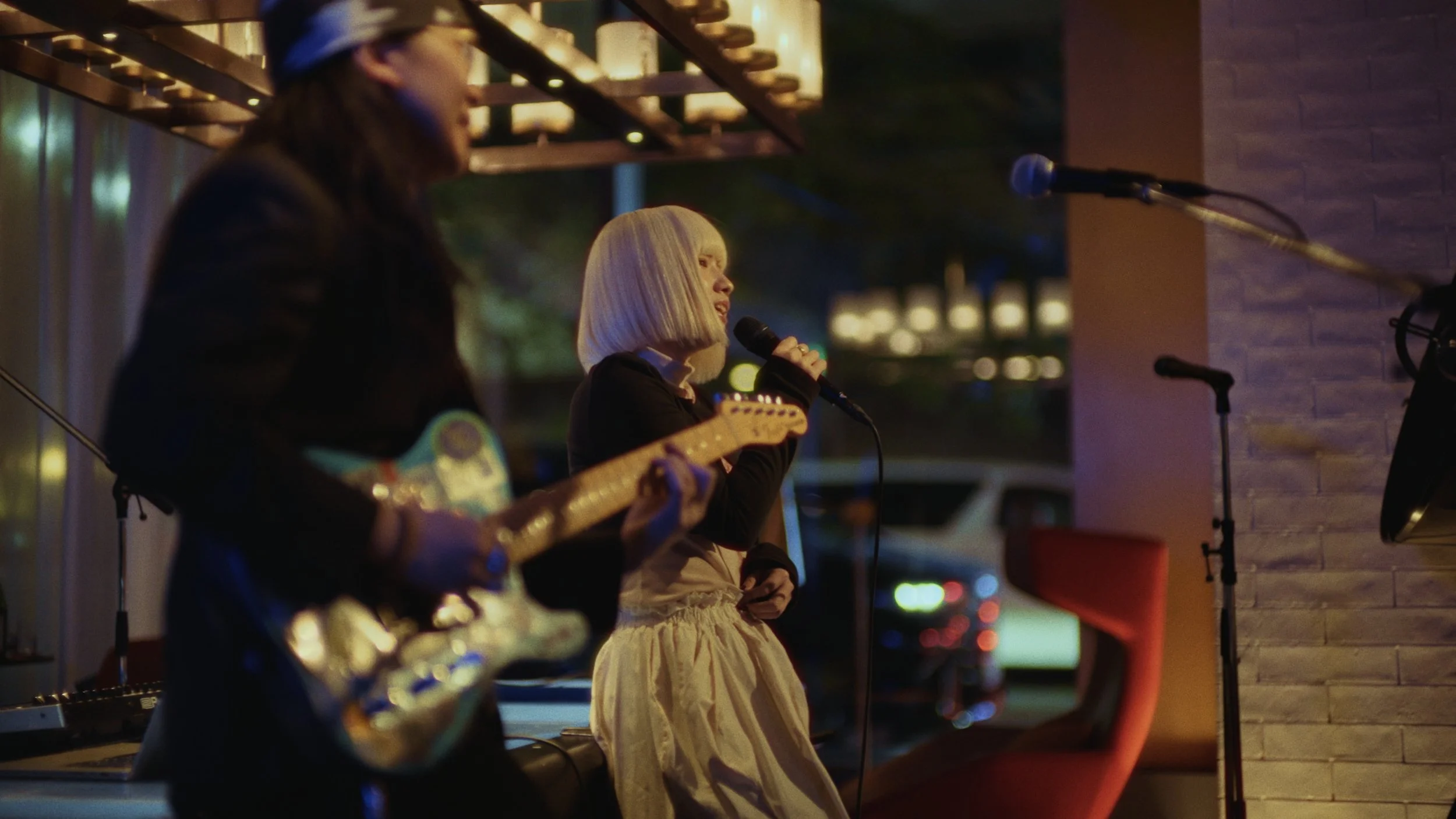 Two women performing on an outdoor stage at night, one playing guitar and the other singing into a microphone, with a brick wall and blurred city lights in the background.