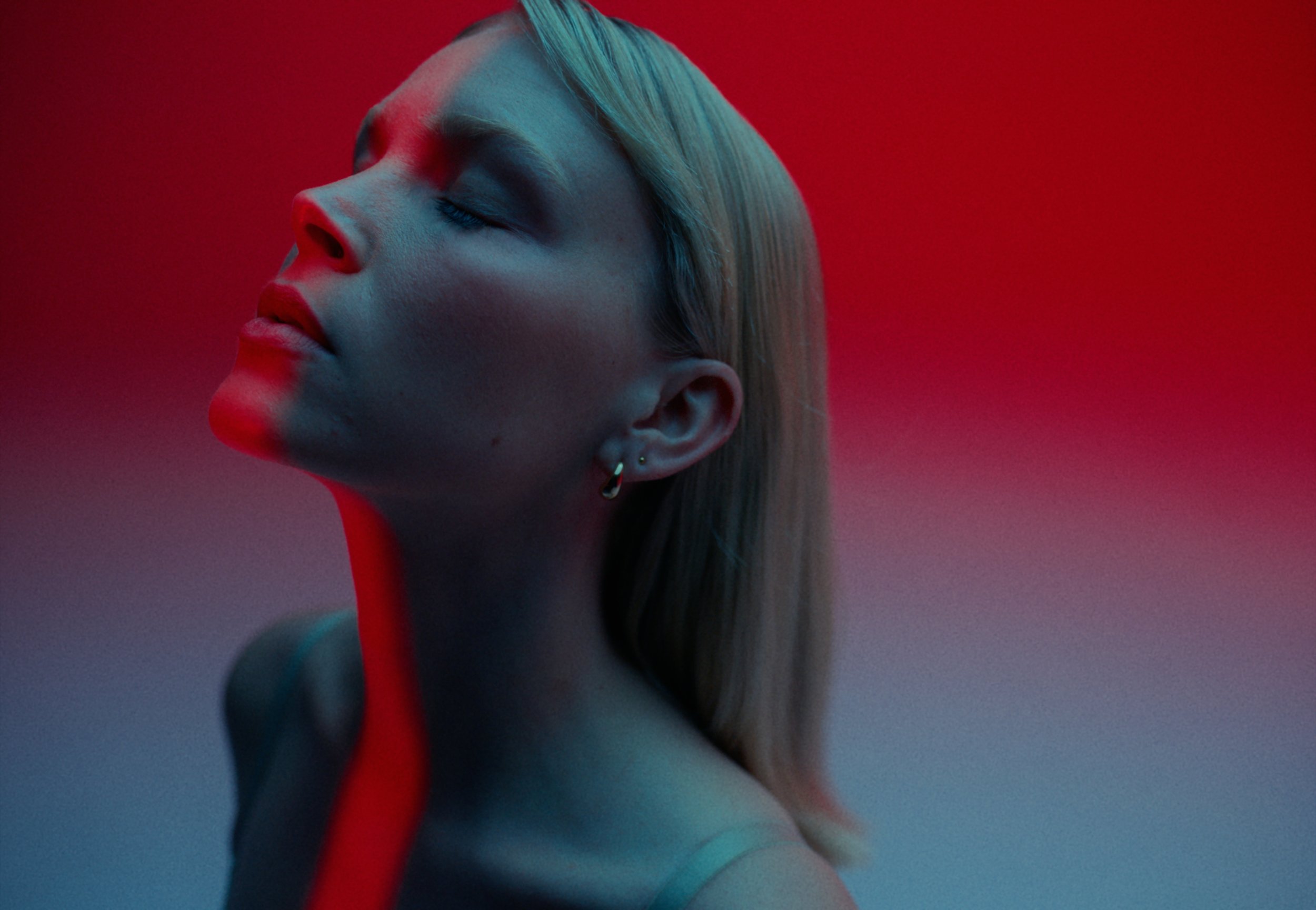A woman with platinum blonde hair, closed eyes, and pierced ears, posed against a red background with soft lighting.