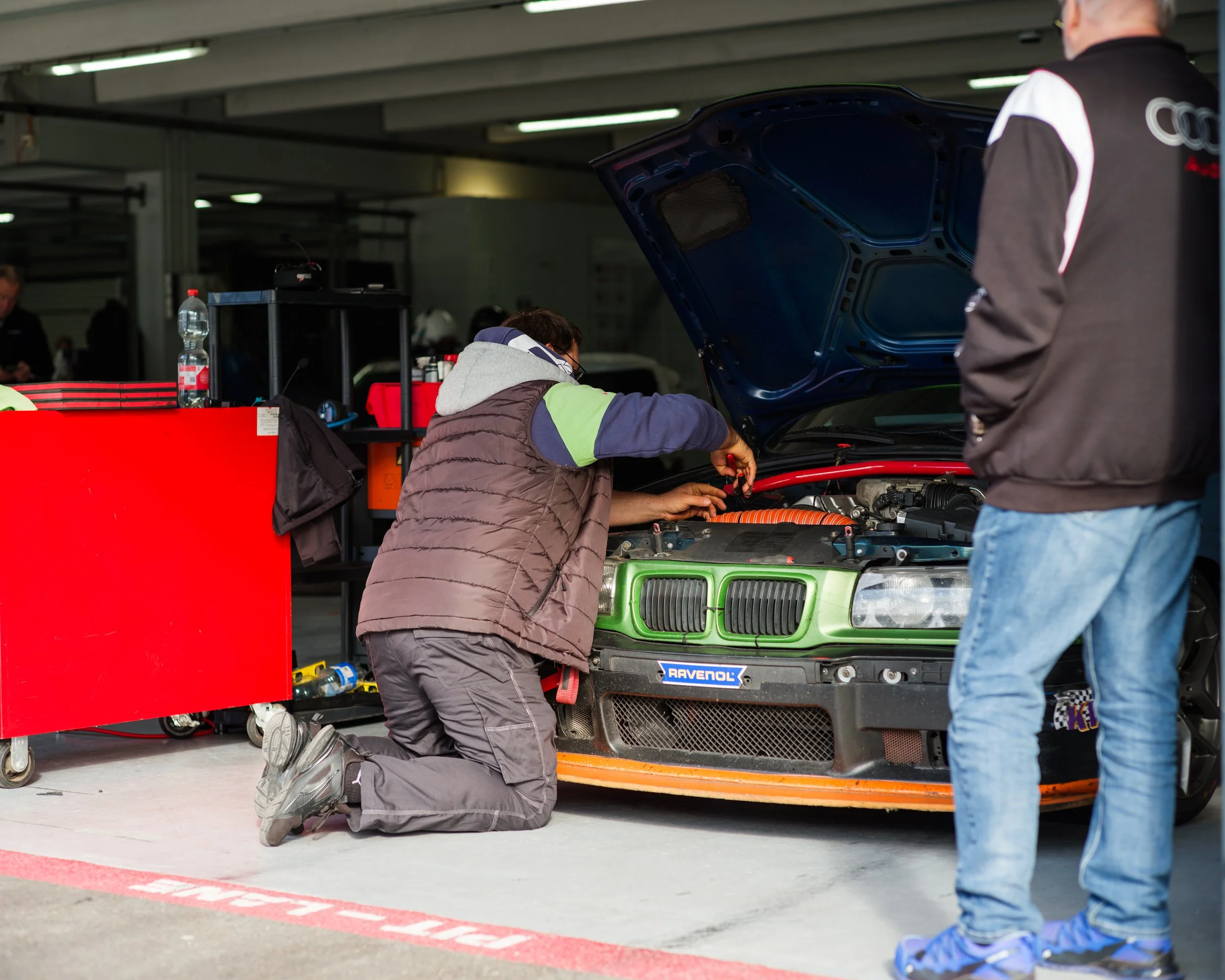 Mechanic kneeling and working on a green race car in a garage, with another person standing nearby and tools and equipment around.