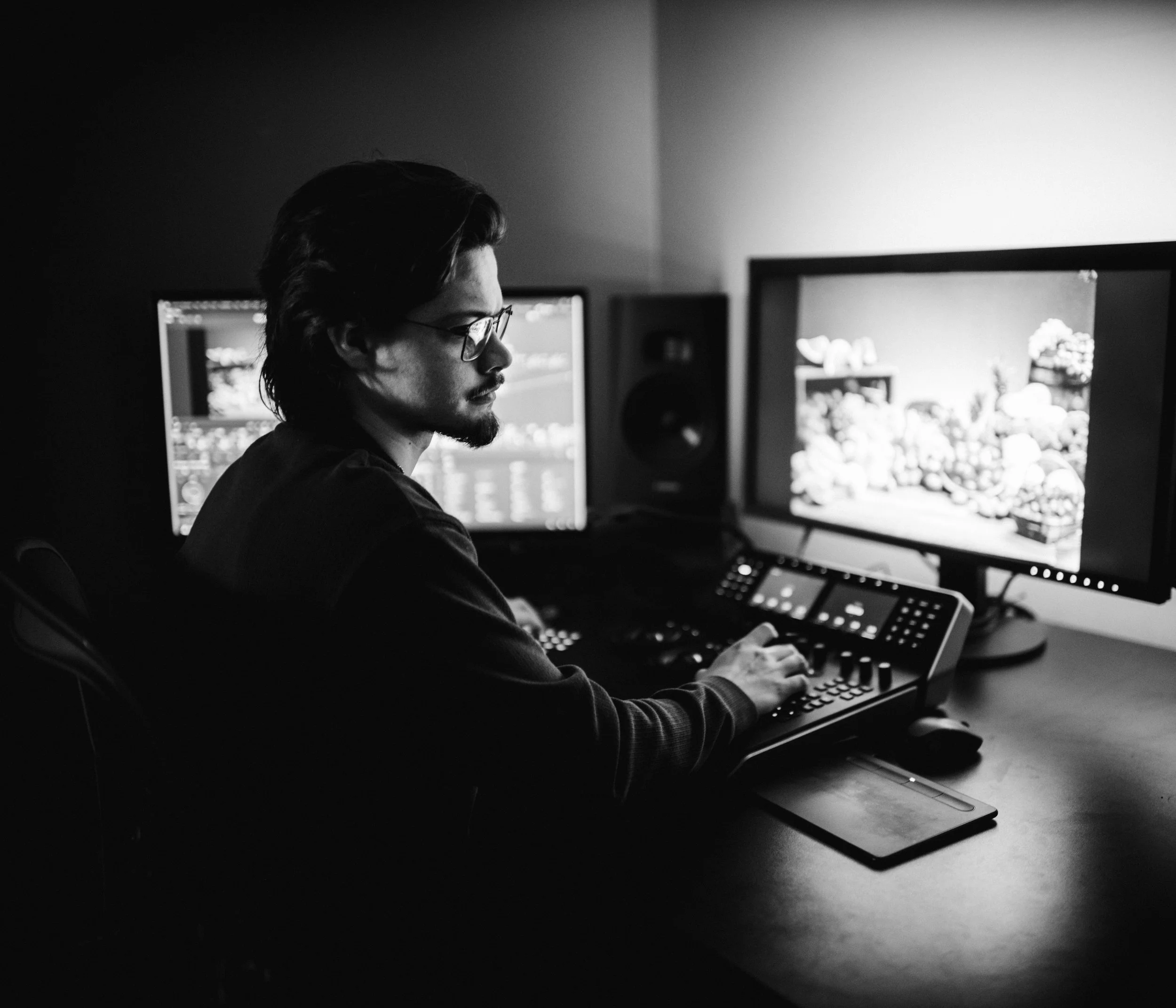 A man with glasses working on a computer in a dimly lit room, with multiple monitors and audio speakers.