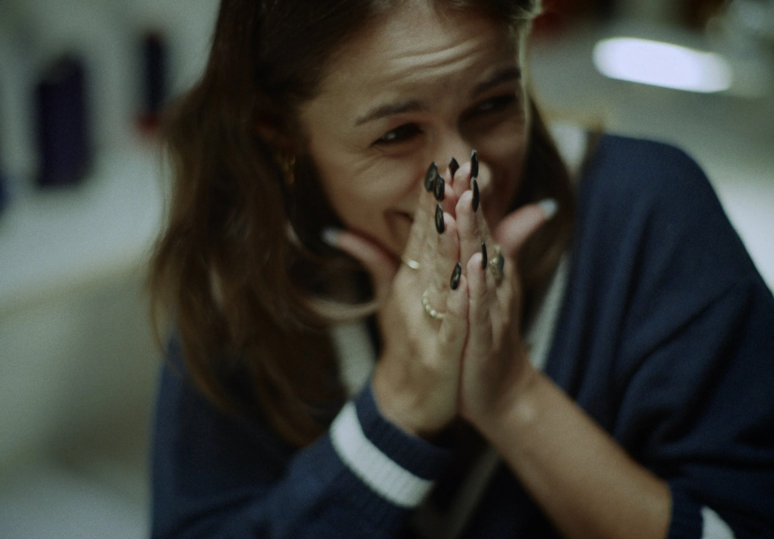 A woman with brown hair tied back, wearing a dark blazer and a white top, covers her face with her hands and appears to be smiling or laughing, in an indoor setting.