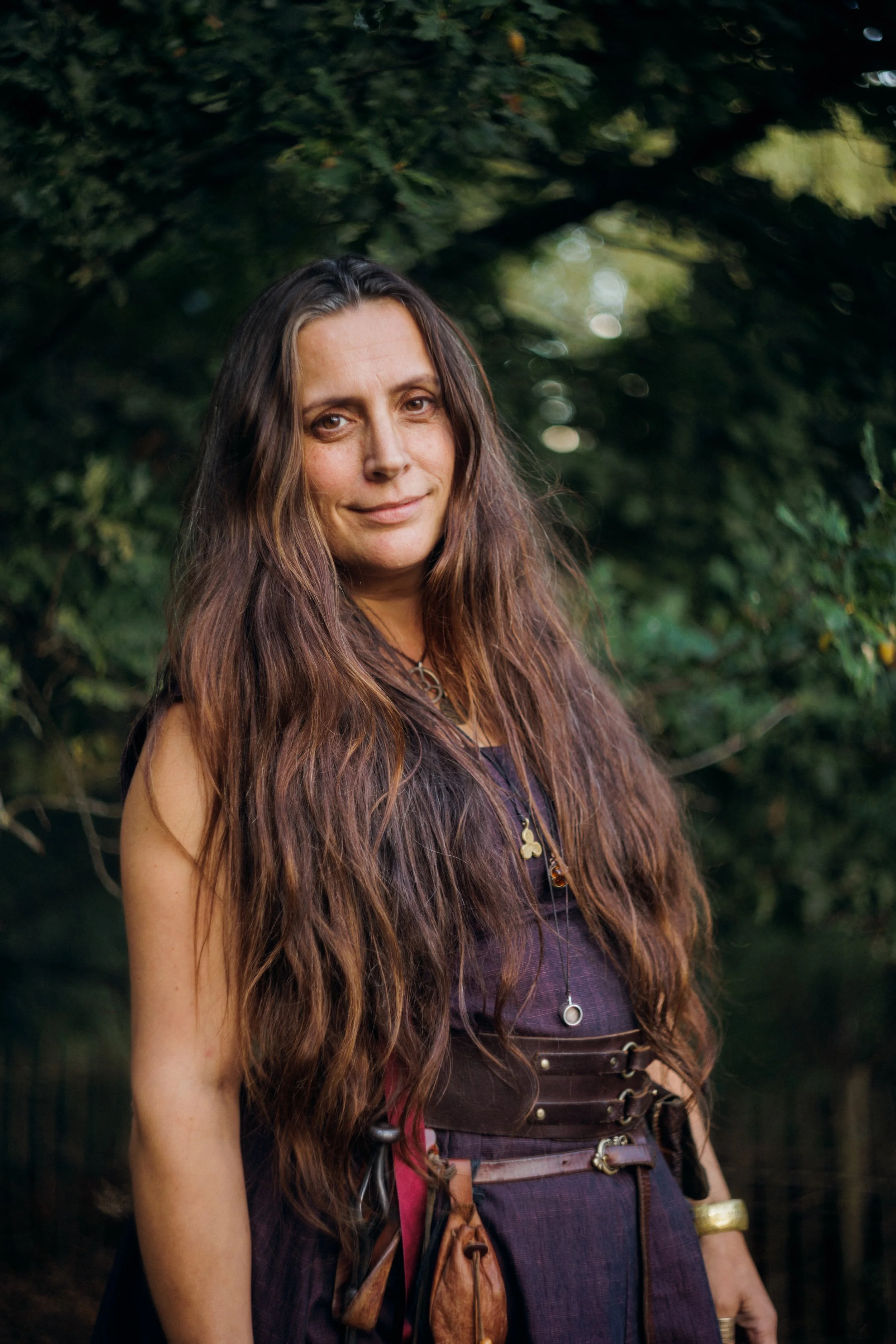 A woman with long brown hair standing outdoors in front of greenery, wearing a purple top, a belt with metal rings, and various jewelry.