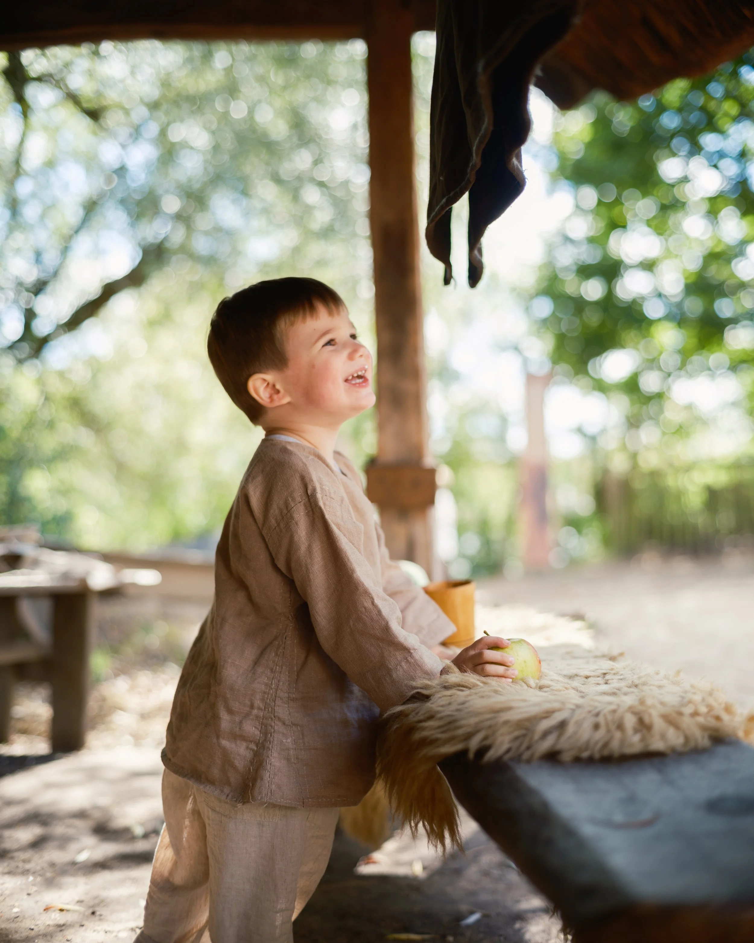 A young boy with brown hair wearing a beige long sleeve shirt and beige pants, smiling while holding a green apple, standing at a wooden table with a furry surface, outdoors under a wooden shelter with blurred green trees in the background.