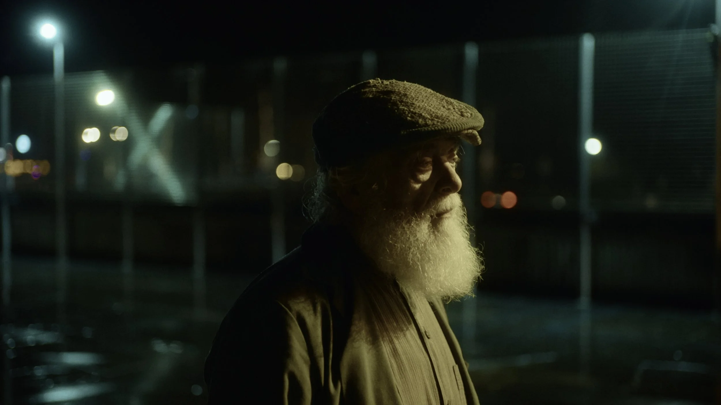 An elderly man with a white beard and mustache wearing a brown cap and brown jacket, standing outdoors at night near a metal fence with lights in the background.