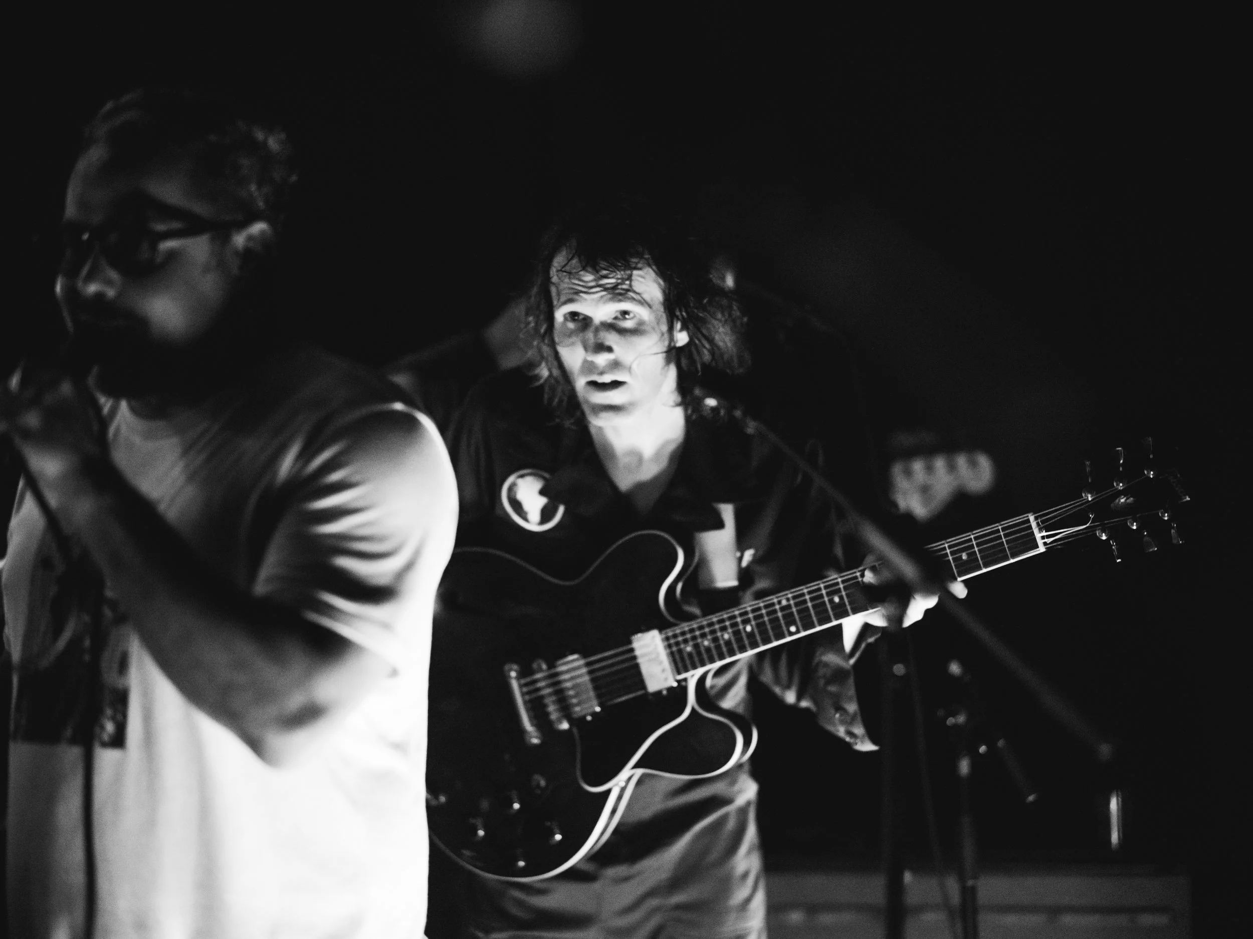 Black and white photo of a person with glasses playing an electric guitar, with another person with long curly hair looking at them, in a dark setting.
